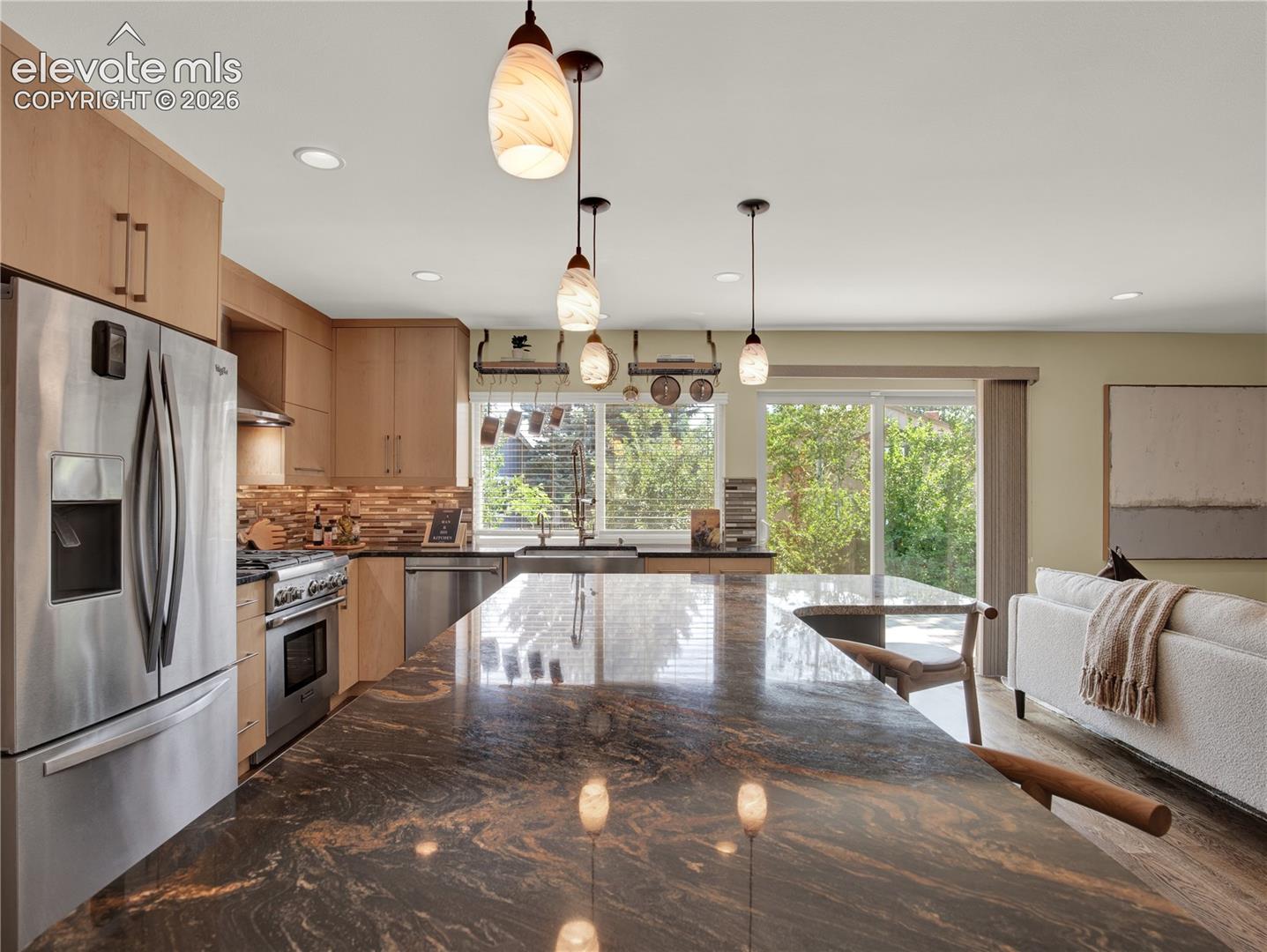Kitchen featuring an expansive stone island, custom wood-finish cabinetry, stainless steel appliances, decorative pendant lighting, and sliding glass doors