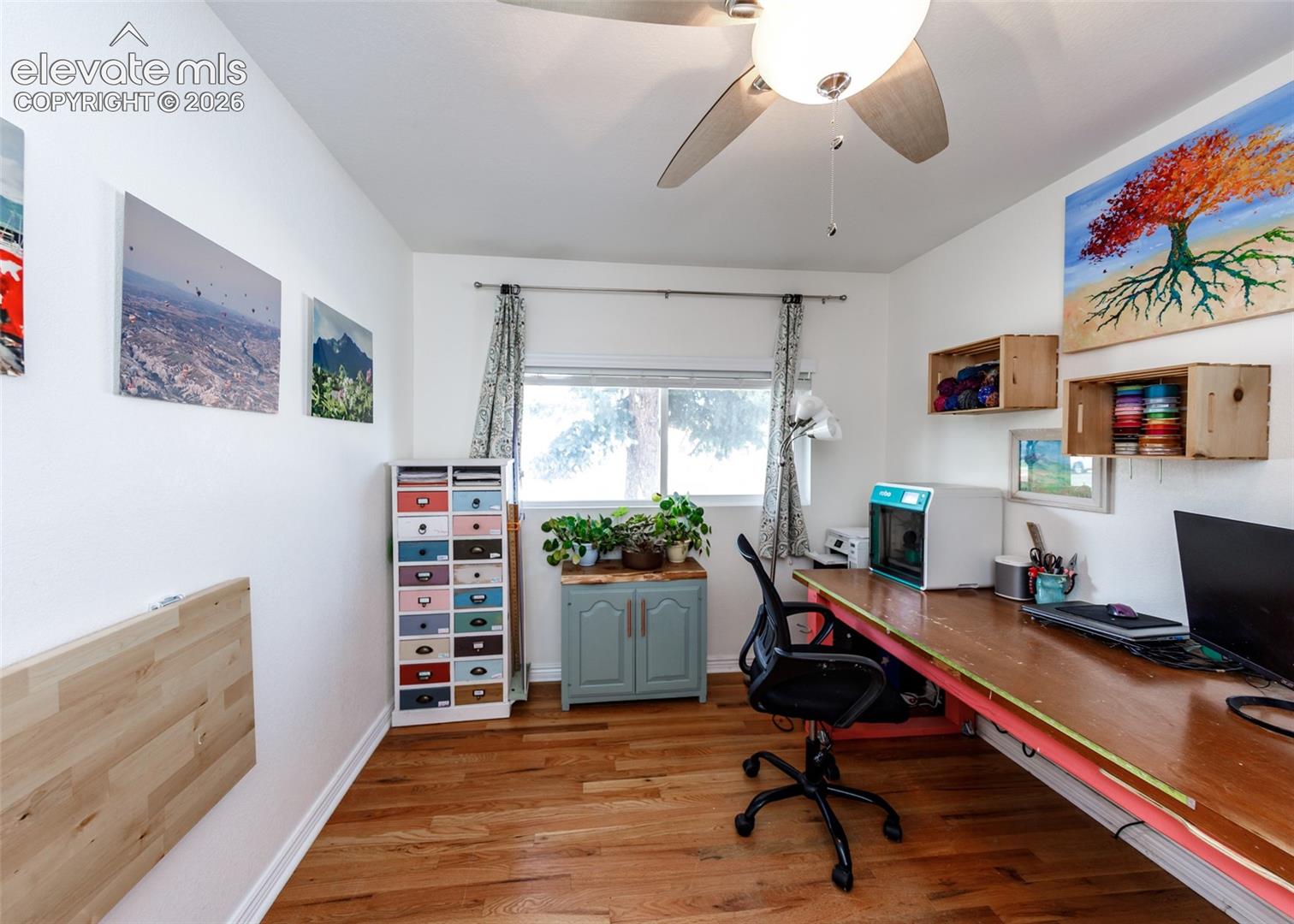 Wood-finish flooring, ceiling fan with light fixture, built-in wall desk, window with roller shades, and built-in shelving