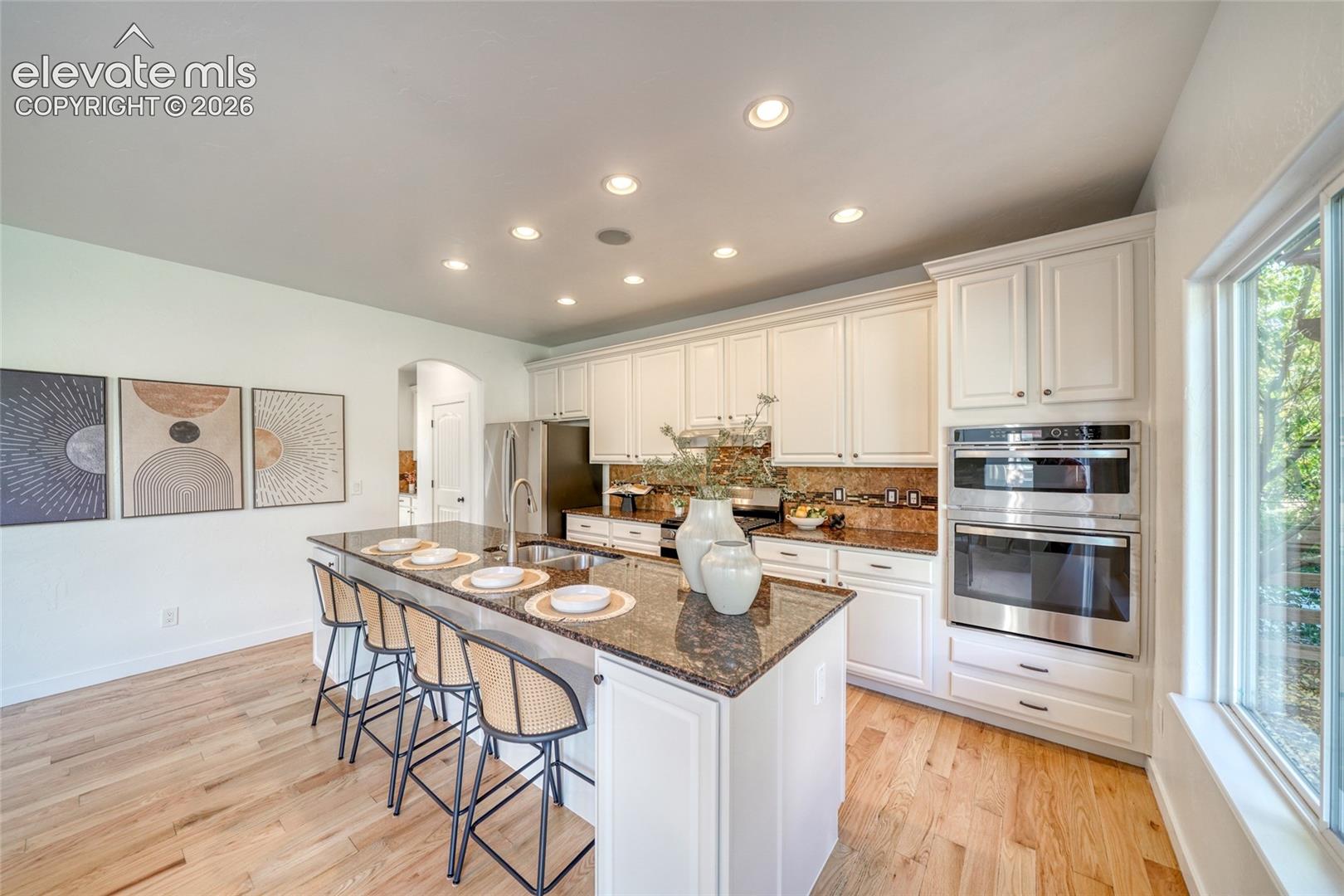 Kitchen featuring a large island with sink, ample white cabinetry, stainless steel built-in double ovens, and wood-finish flooring