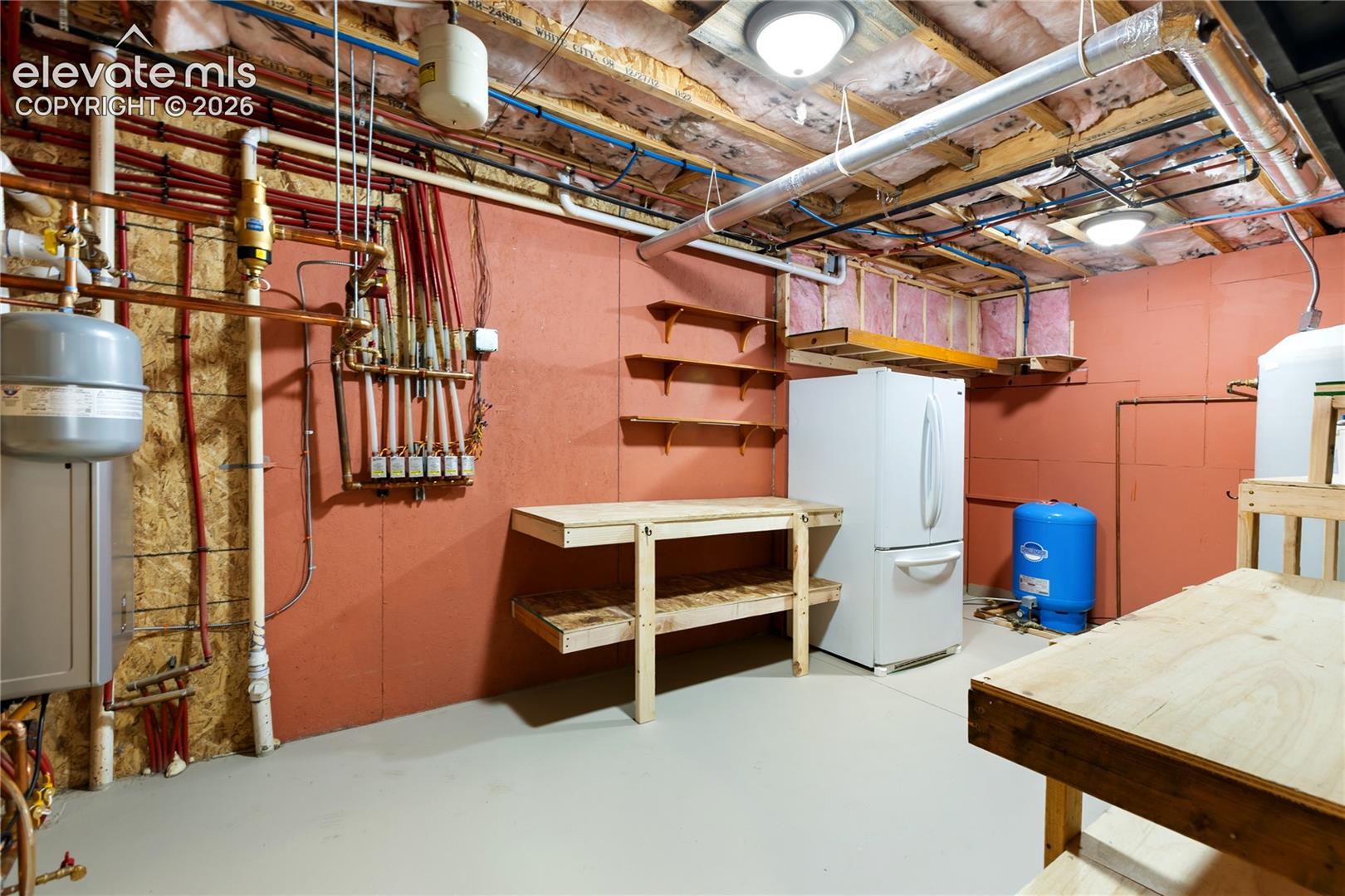 Utility area featuring an exposed plumbing manifold, built-in wooden shelving, and a pressure tank