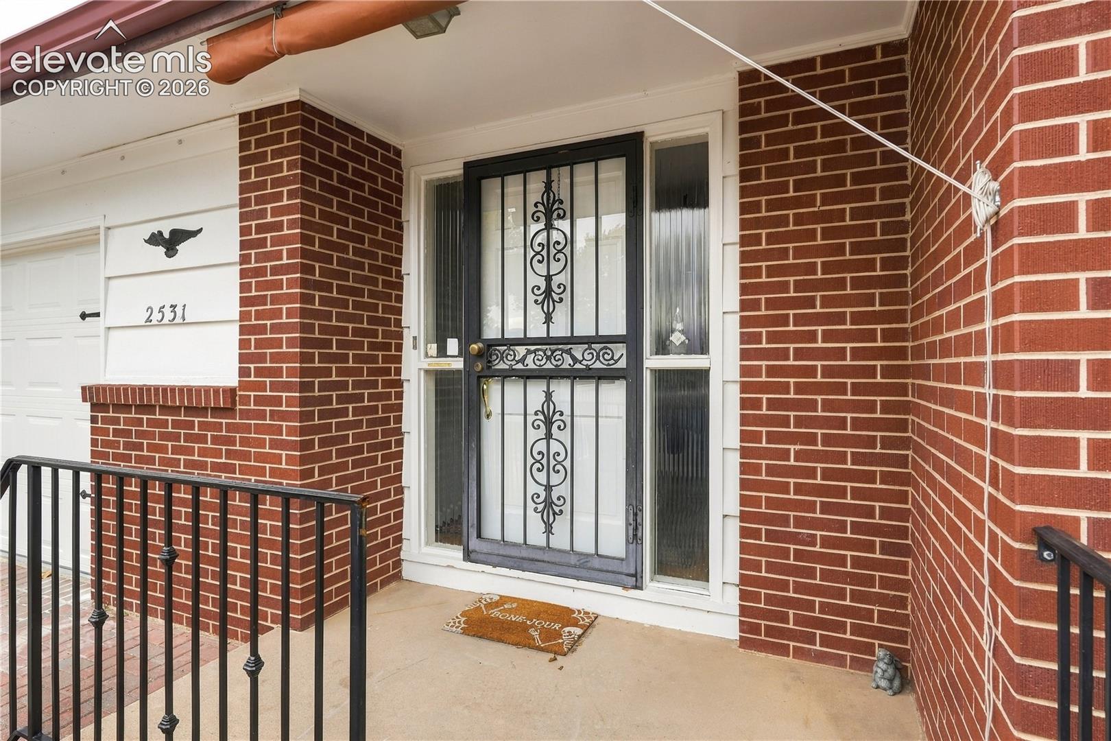 Red brick exterior featuring a covered entry, decorative metal security door, vertical sidelight window, and black metal handrailing