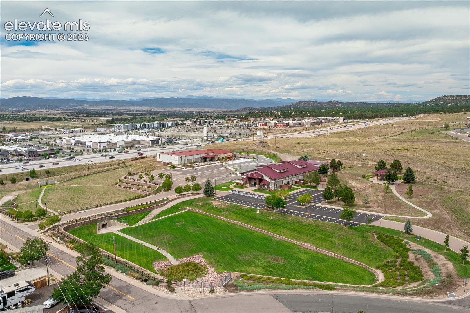 Aerial perspective of a multi-structure site featuring expansive open green space, paved parking areas, masonry retaining walls, and a prominent mountain range backdrop