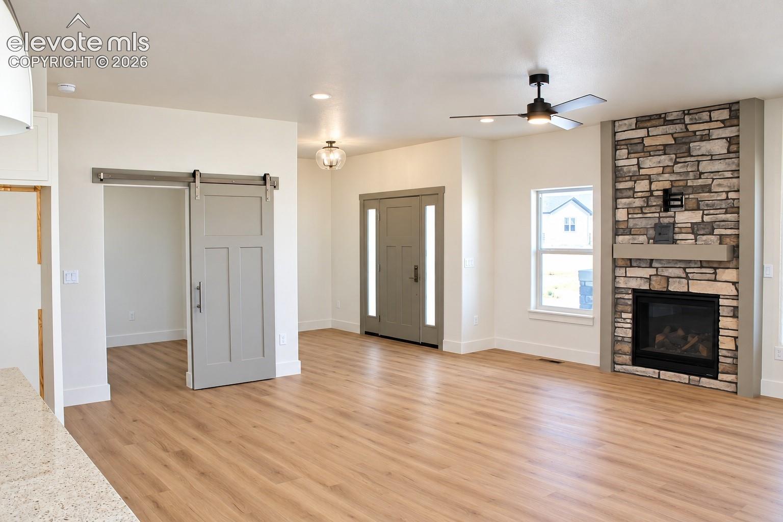 Wood-finish flooring throughout, stone-clad corner fireplace with a built-in mantel, ceiling fan with integrated lighting, and a modern barn door