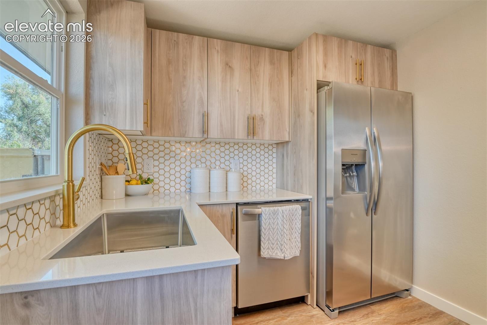 Light wood-finish cabinetry, stainless steel appliances, white hexagon tile backsplash, light-toned countertop, and a gold-finish kitchen faucet