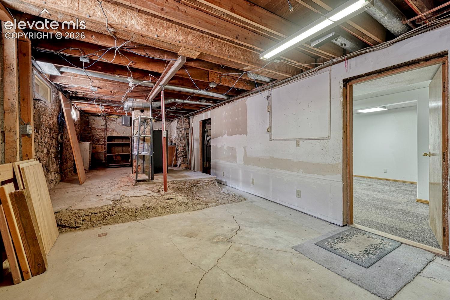 Exposed wood joists, concrete flooring, stone foundation wall, and a separate room with carpeted flooring