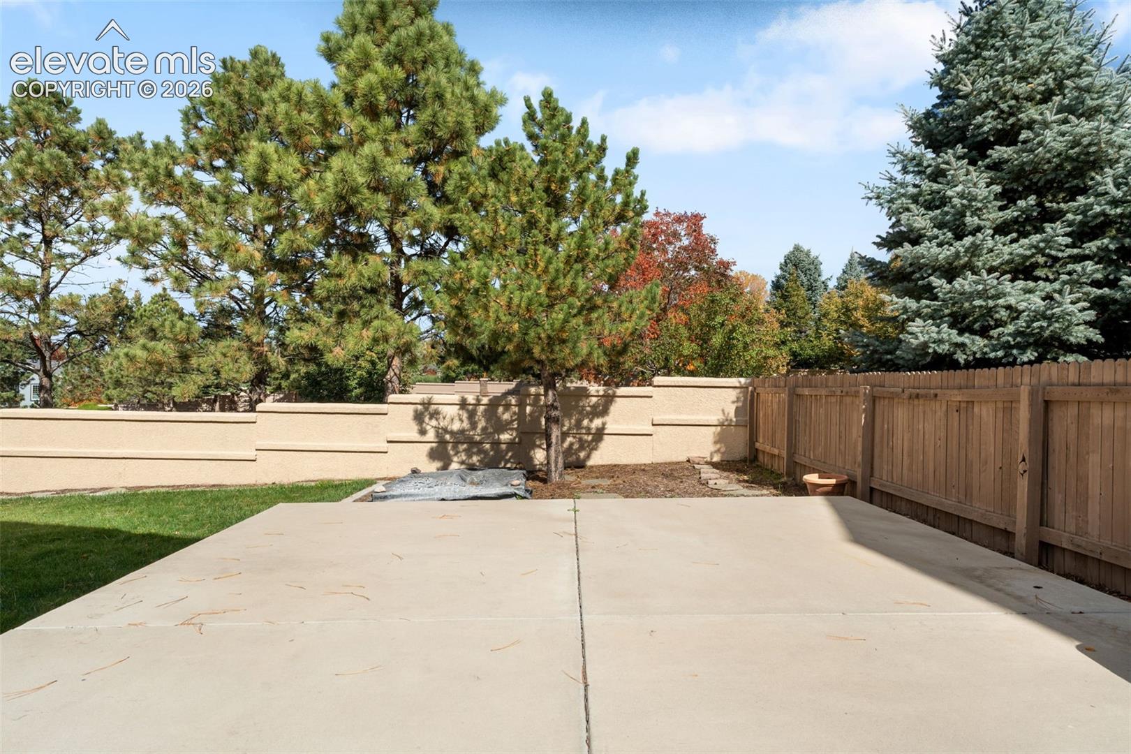 Concrete patio featuring a perimeter of wood fencing and stucco wall boundaries, complemented by a manicured lawn and mature coniferous trees
