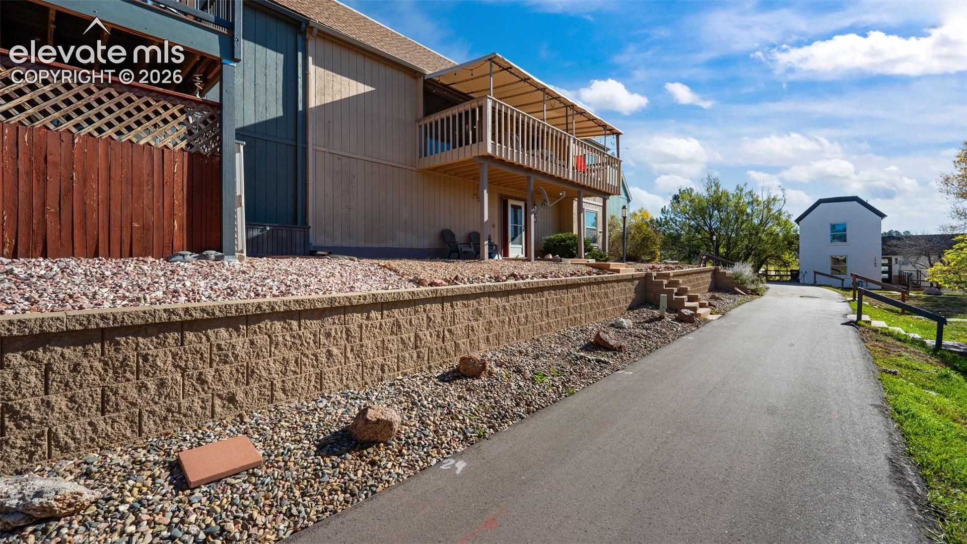 Exterior features including vertical wood-panel siding, a covered second-story deck with wooden railings, and a concrete block retaining wall encompassing a gravel-landscaped yard area
