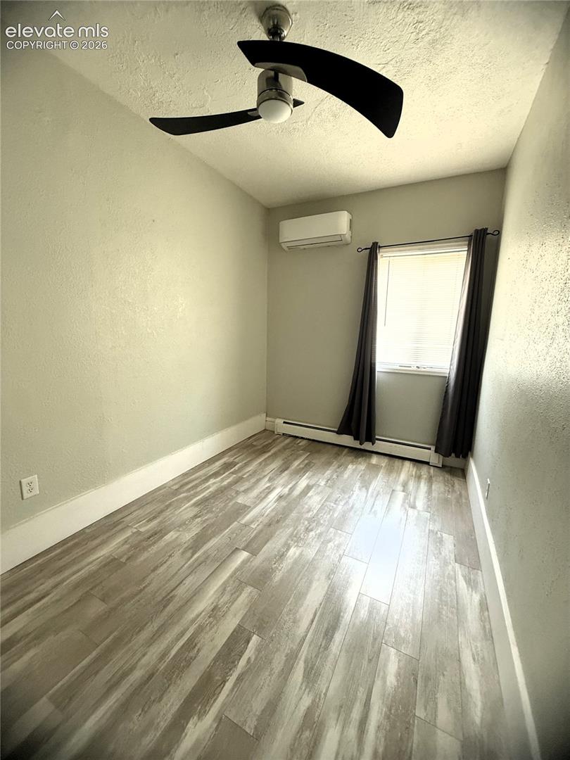 Wood-finish flooring, wall-mounted ductless mini-split unit, ceiling fan with light fixture, and window with horizontal blinds