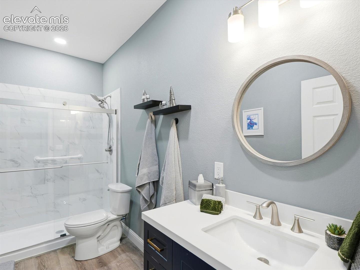 Basement bathroom Vanity featuring a solid-surface countertop and undermount sink, glass-enclosed shower with tiled surround, wood-finish flooring, and circular mirror with metallic frame