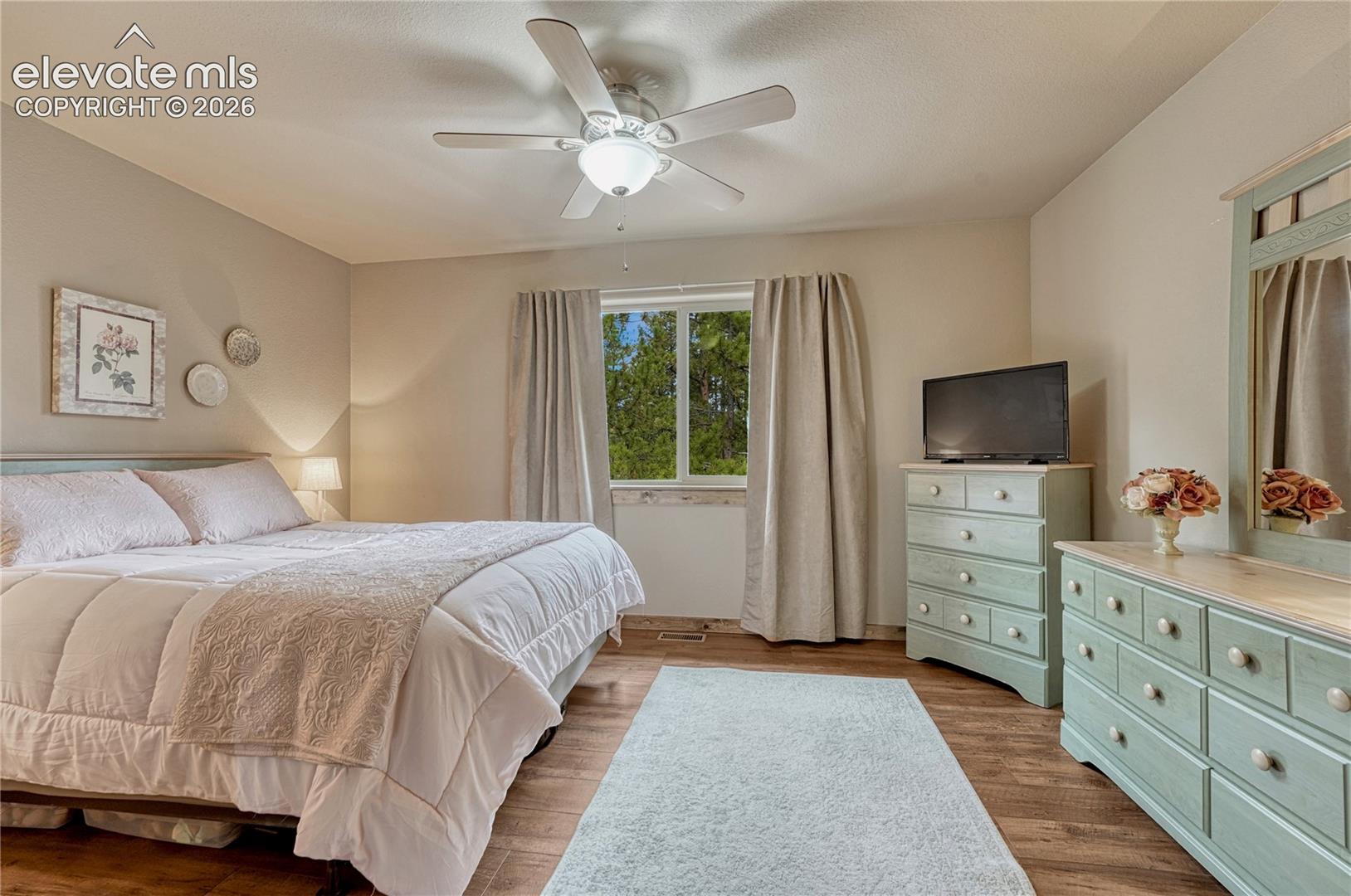 Wood-finish flooring, ceiling fan with integrated lighting, and window featuring exterior tree-line scenery