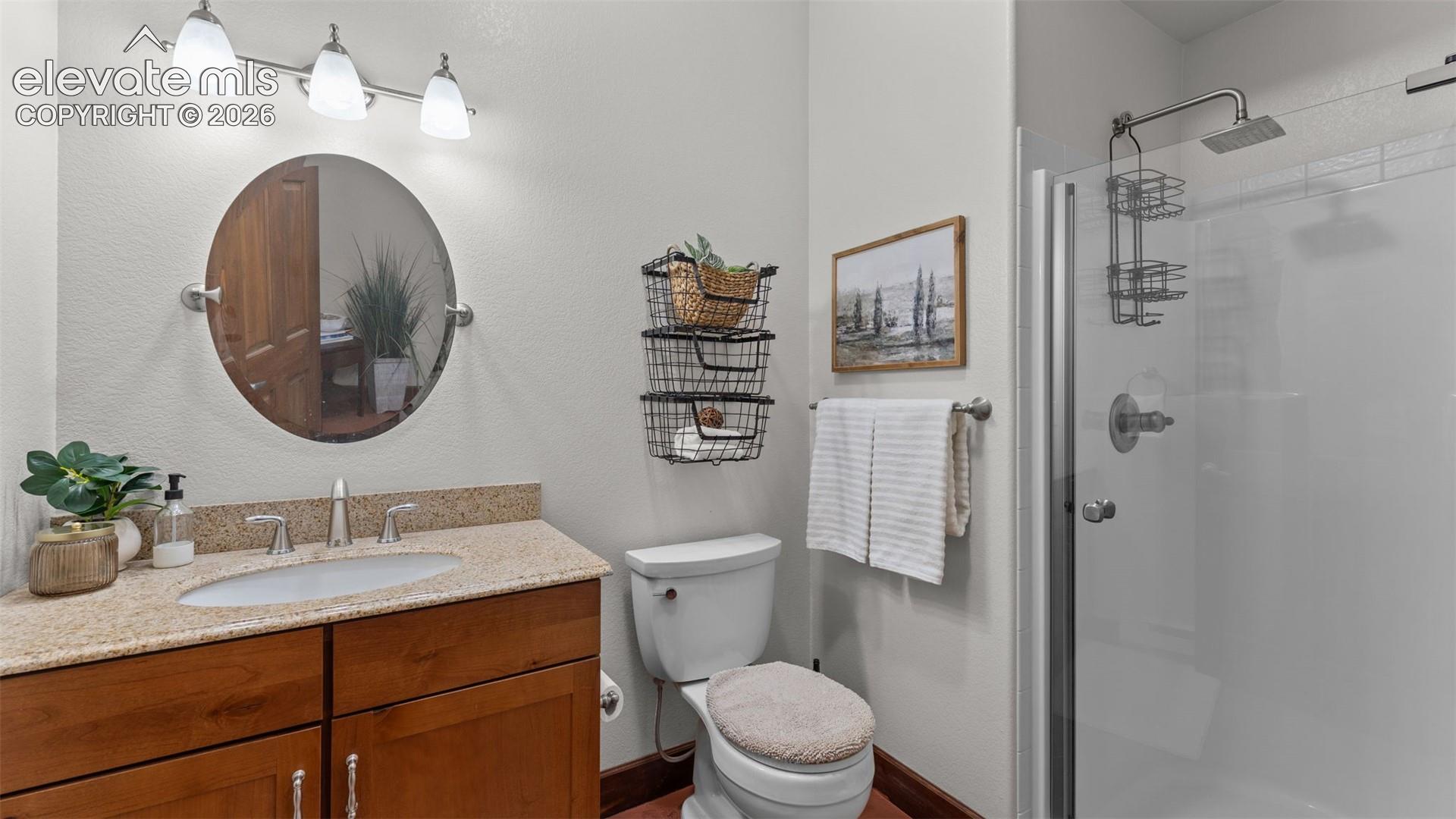 Bathroom featuring a wood-tone vanity, solid surface countertop with integrated sink, circular wall-mounted mirror, three-light vanity fixture, and a glass-enclosed shower with a rainfall showerhead