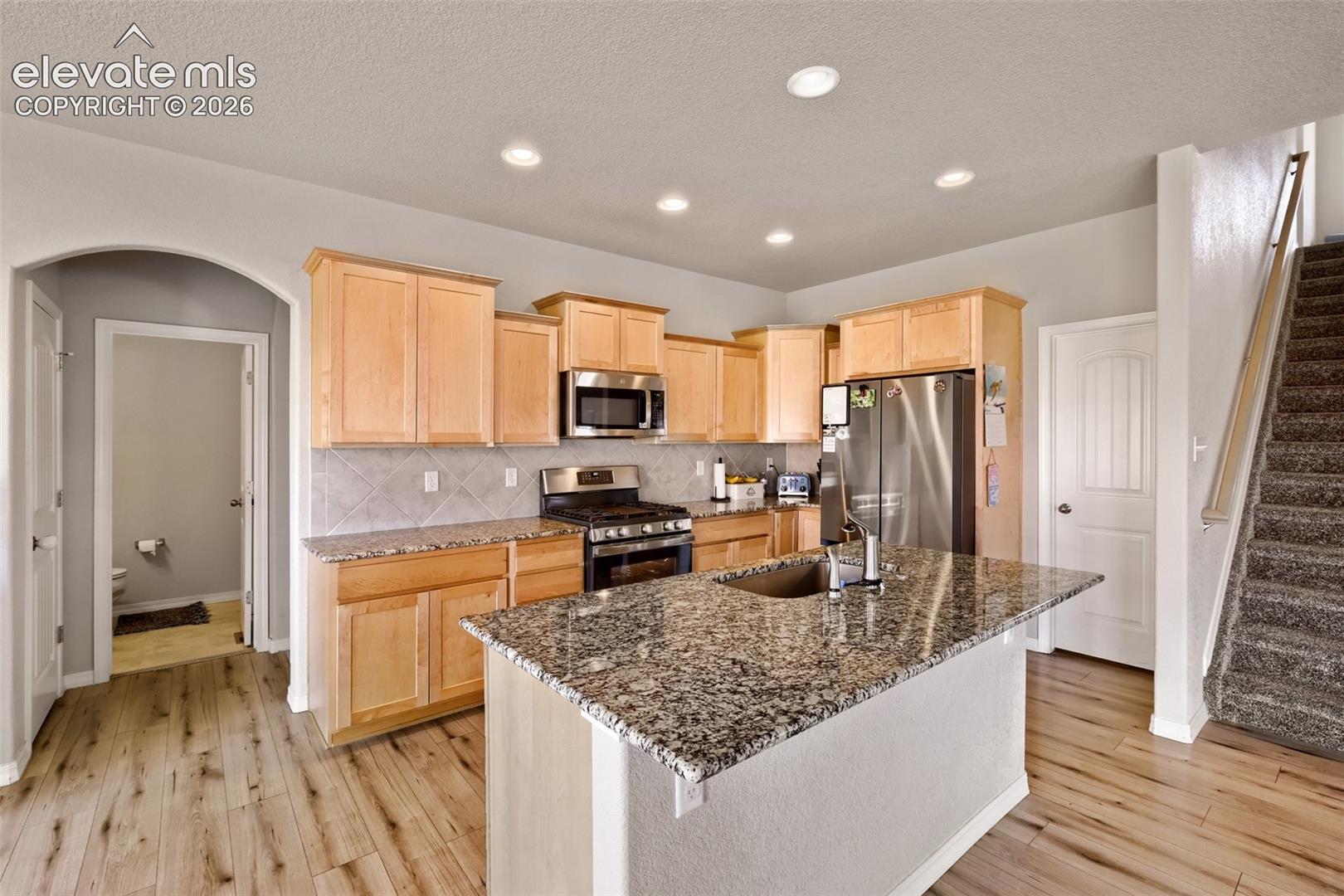 Kitchen featuring wood-finish flooring, a central island with an undermount sink and stone countertop, stainless steel appliances, natural wood cabinetry, and recessed lighting