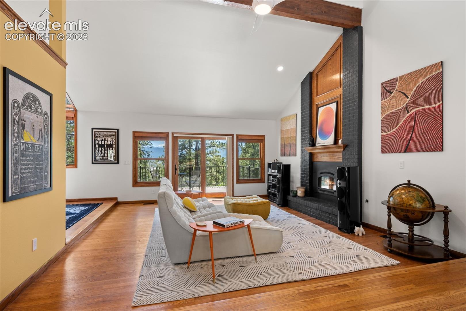 Vaulted living space featuring a black brick fireplace with a wood mantle, hardwood flooring, wood-frame windows, and exposed wood beams