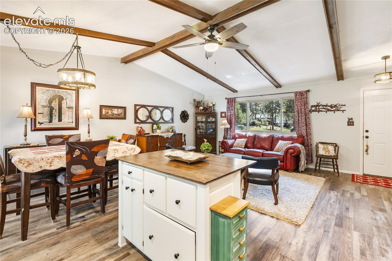 Vaulted ceiling with exposed wood beams, wood-finish flooring, white kitchen island with wood countertop, front entry door, and large window