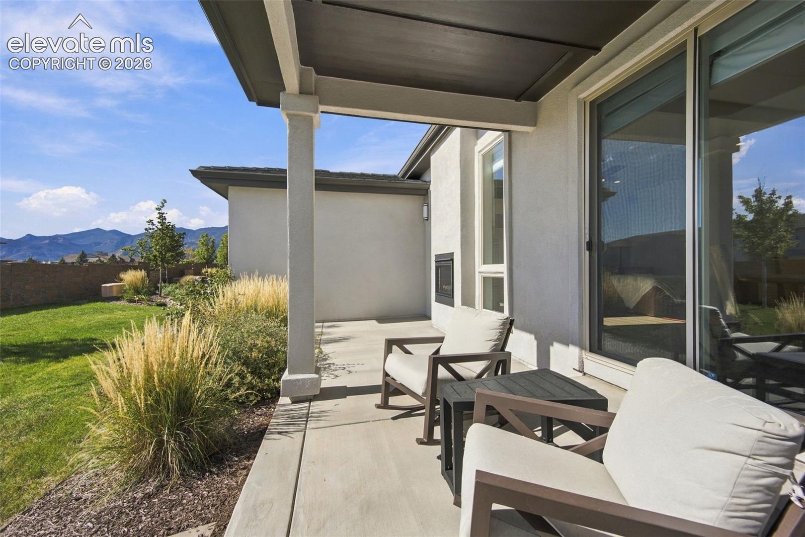 Covered concrete patio featuring an integrated outdoor fireplace, square support columns, and sliding glass door access, overlooking a landscaped backyard with mountain scenery