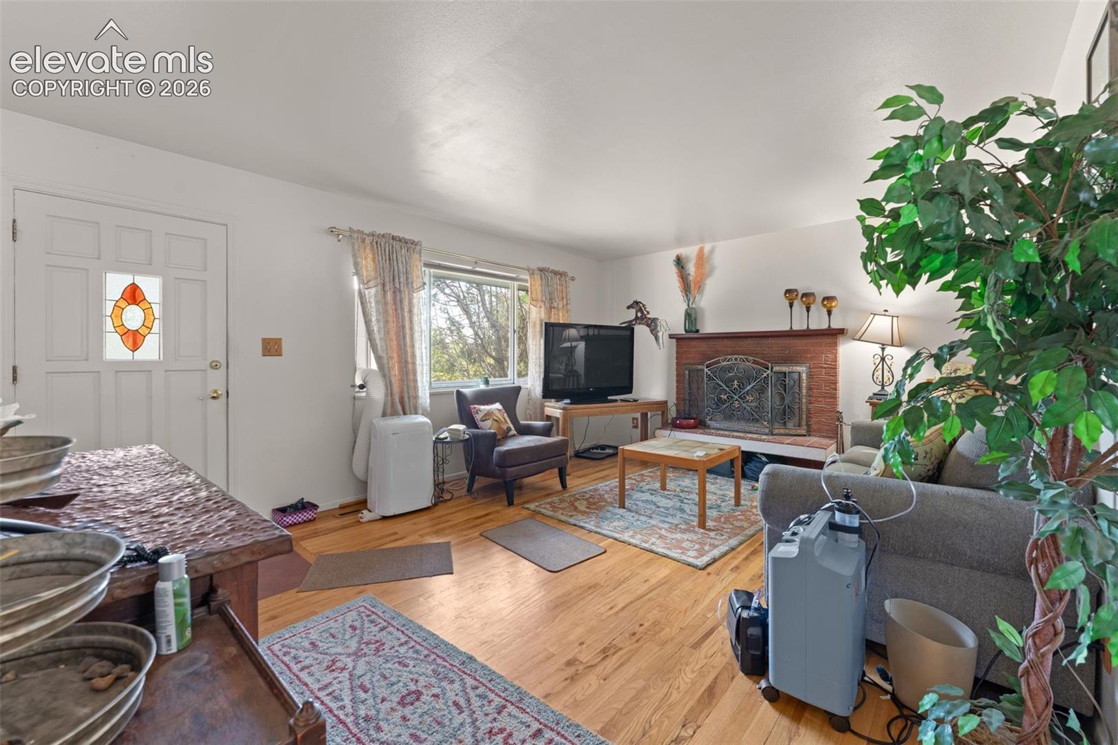 Hardwood-finish flooring, a white paneled entry door with stained glass insert, a brick fireplace with a metal screen, and white walls