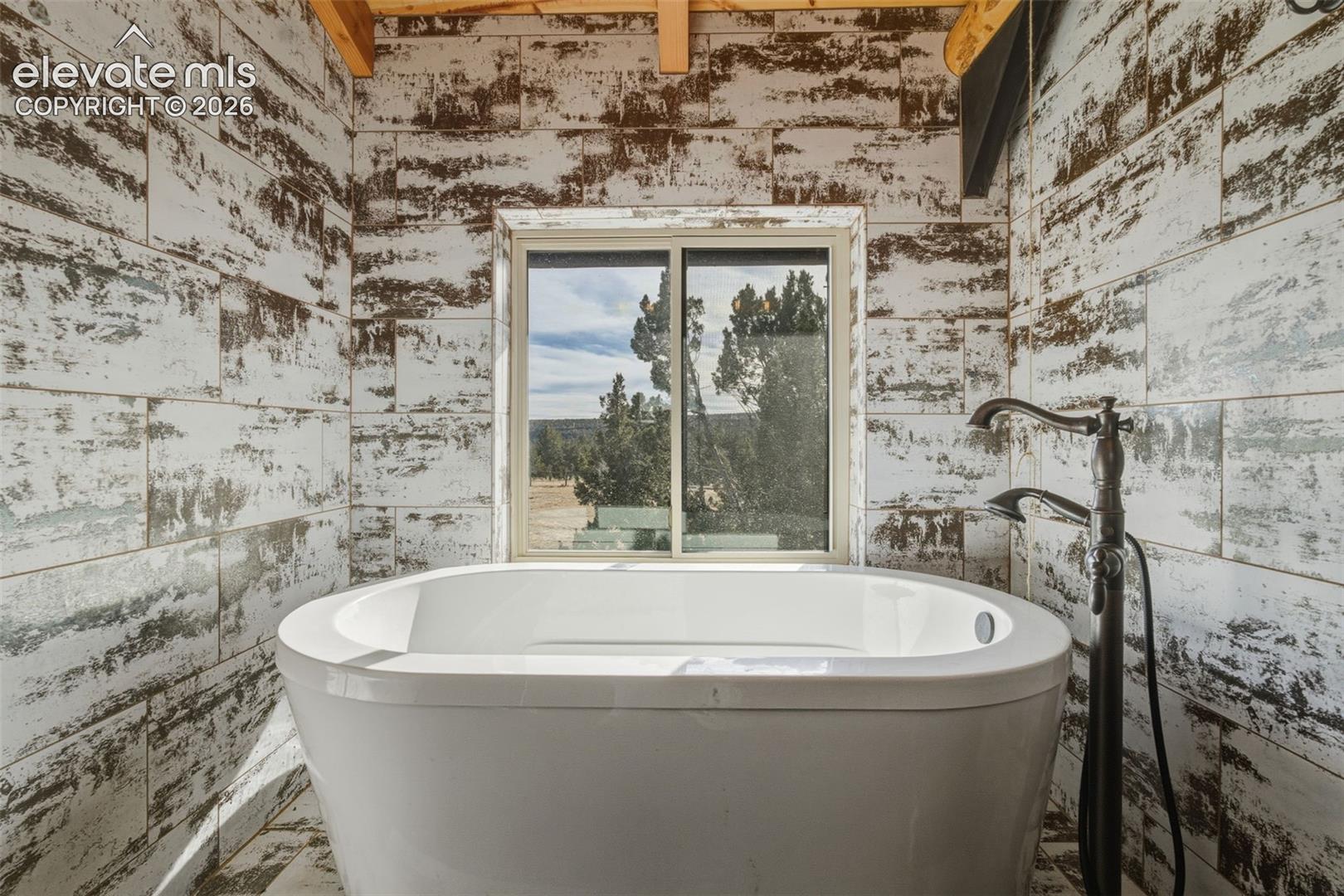 Freestanding soaking tub with a floor-mounted bath filler, wood-finish wall tile, and exposed wood ceiling beams