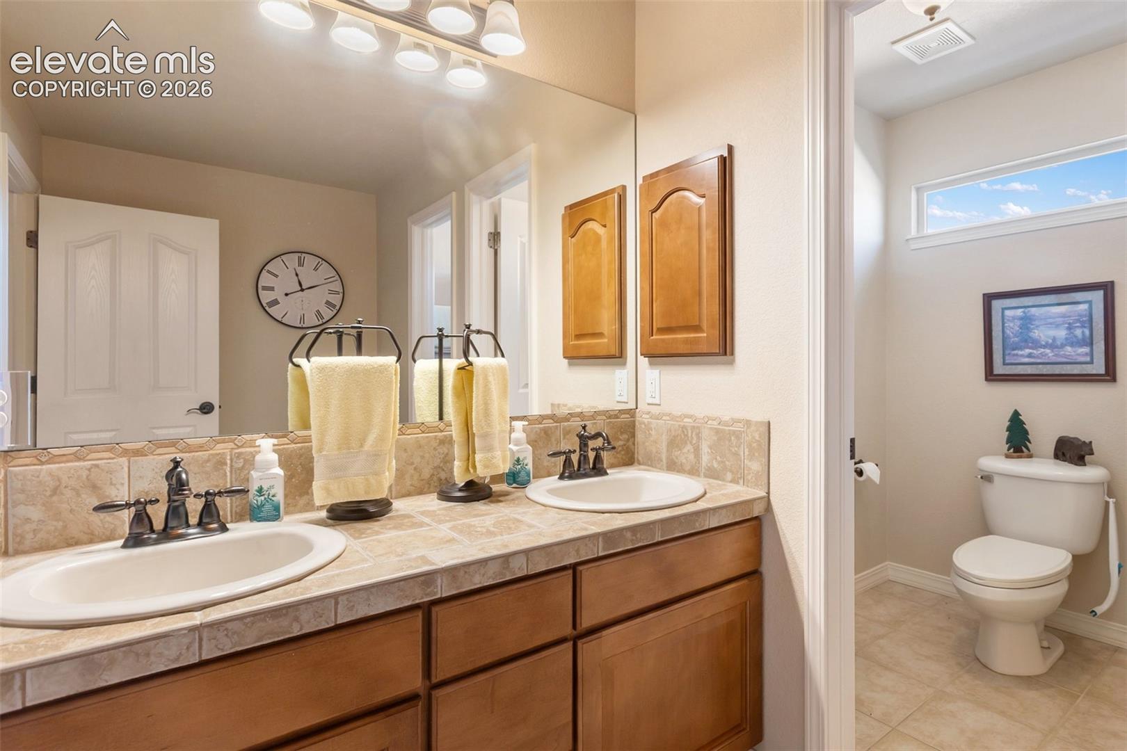Dual-sink vanity with tile countertop and backsplash, wood cabinetry, large vanity mirror with multi-light fixture, and water closet featuring a horizontal transom window In Primary Bathroom