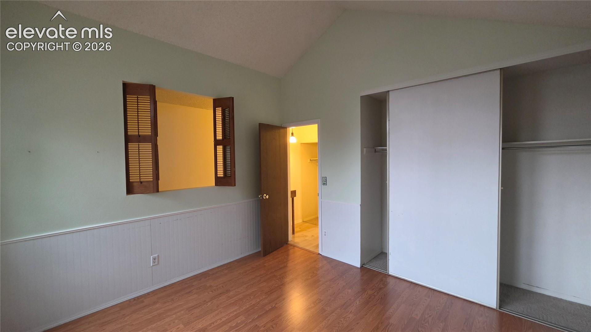 Vaulted ceiling, wood-finish flooring, half-wall paneling, built-in closet with sliding doors, and louvered pass-through window