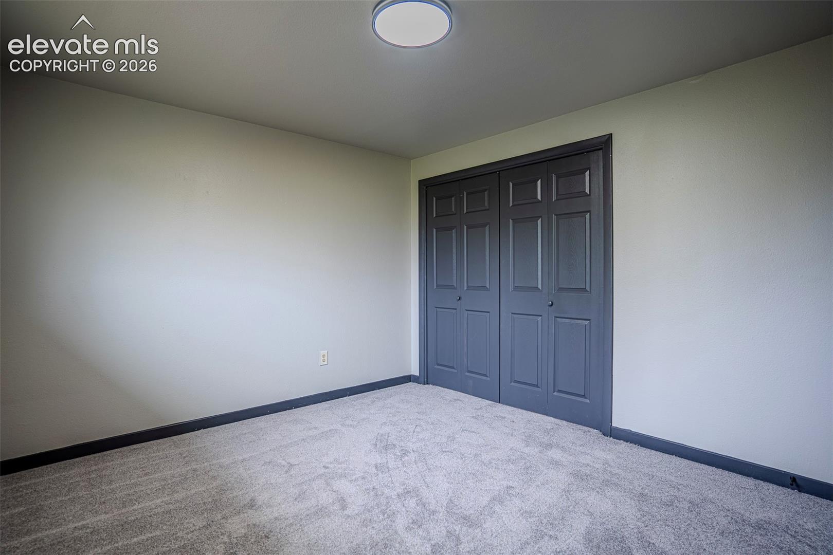 Neutral-toned interior space featuring wall-to-wall carpeting, dual-paneled closet doors with dark-tone trim, and a low-profile flush-mount ceiling light fixture