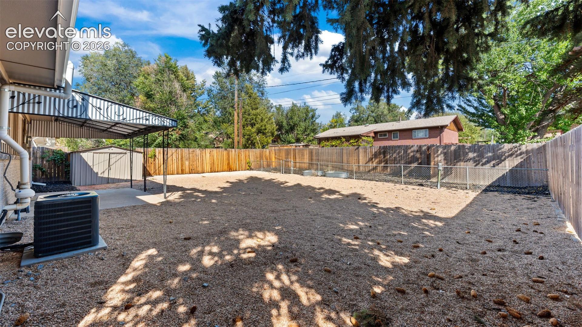 Expansive outdoor area featuring a gravel surface, a covered patio, a storage shed, a wood fence, and mature trees