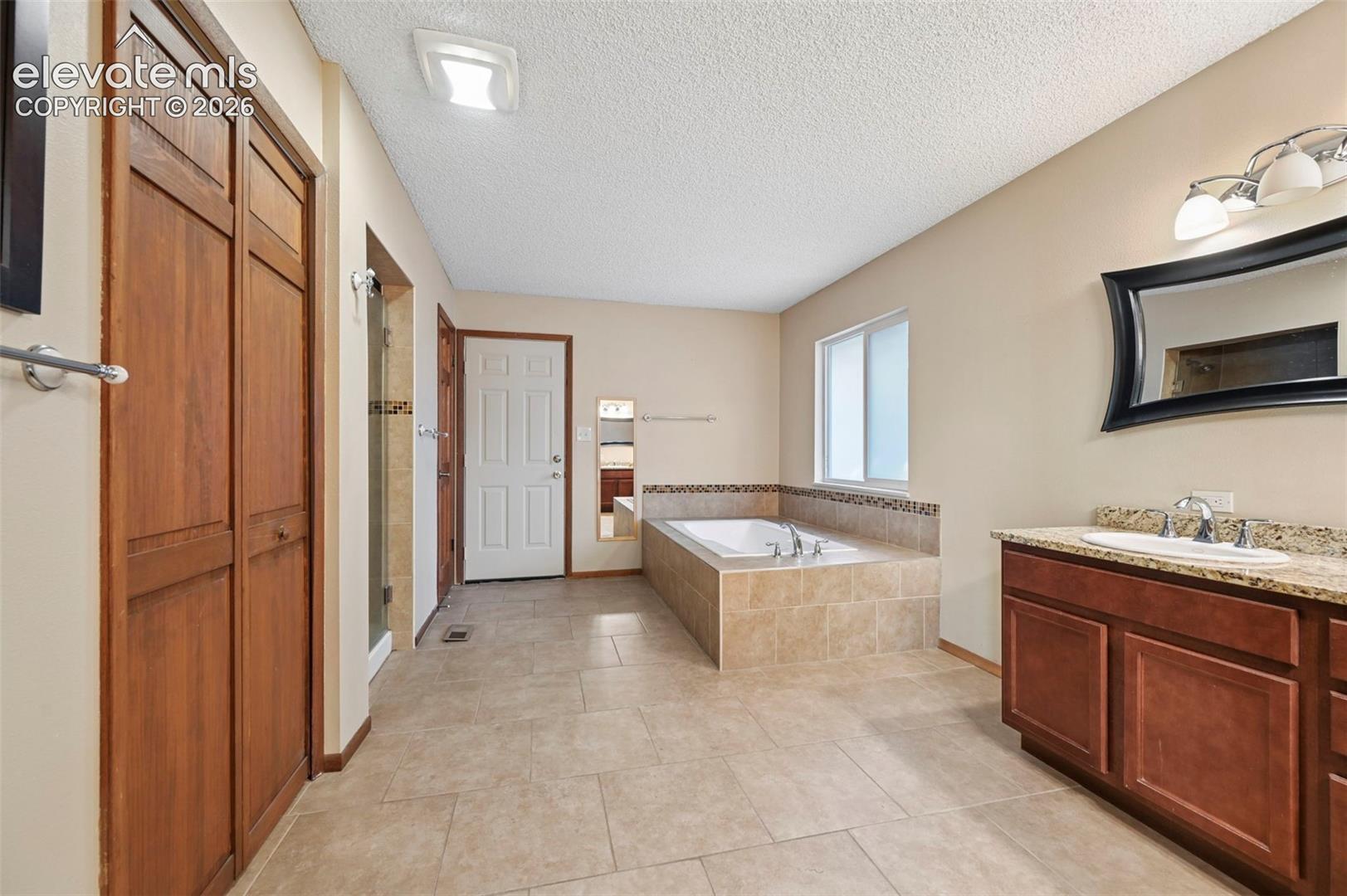 Bathroom featuring a tile-surround soaking tub, vanity with stone-finish countertop, dual-door wood-finish cabinetry, and ceramic tile flooring