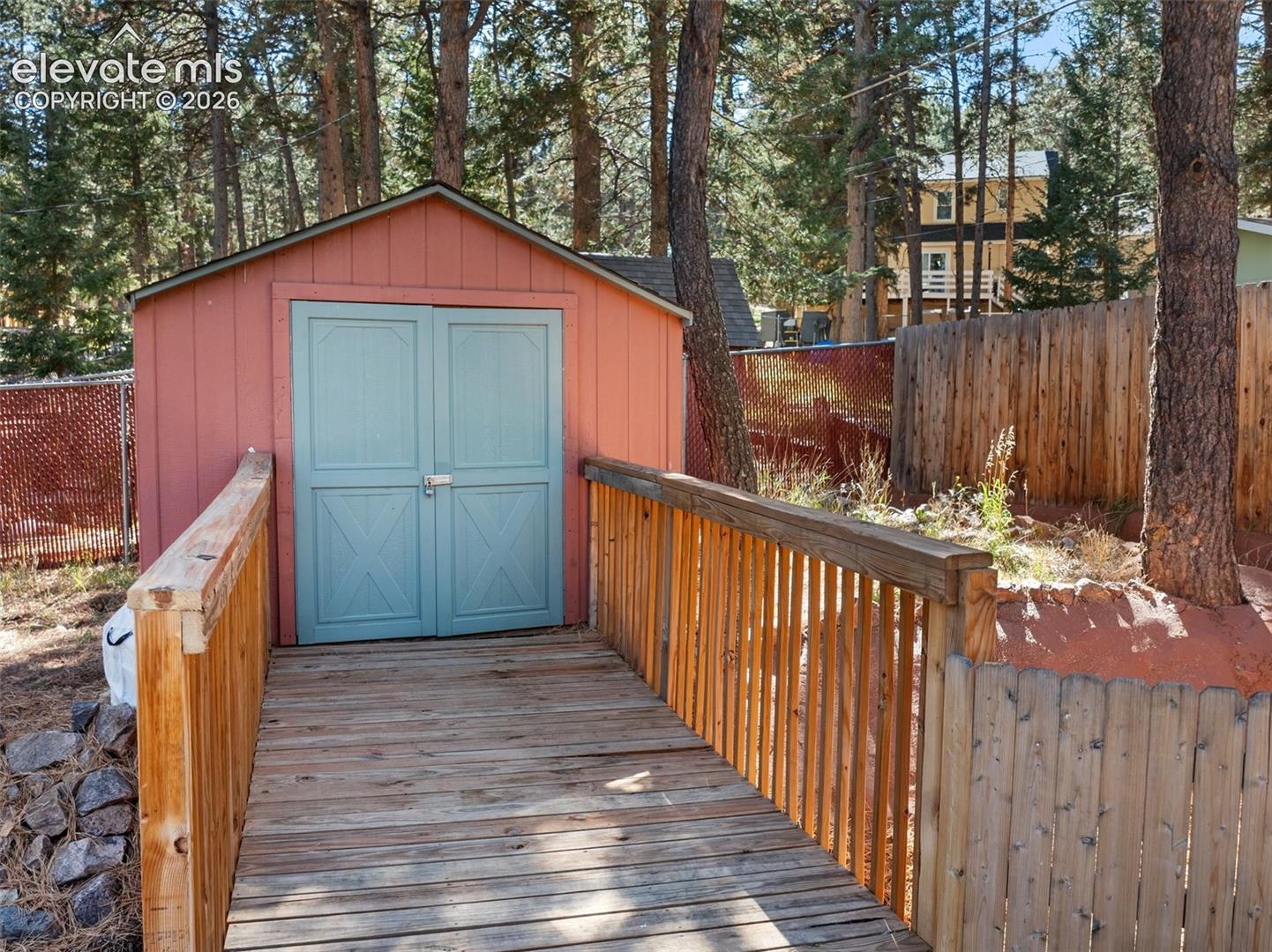 Storage shed with double doors featuring an elevated wooden bridge entrance with sloped railings, situated on a lot with mature pine trees and wood-fencing enclosures