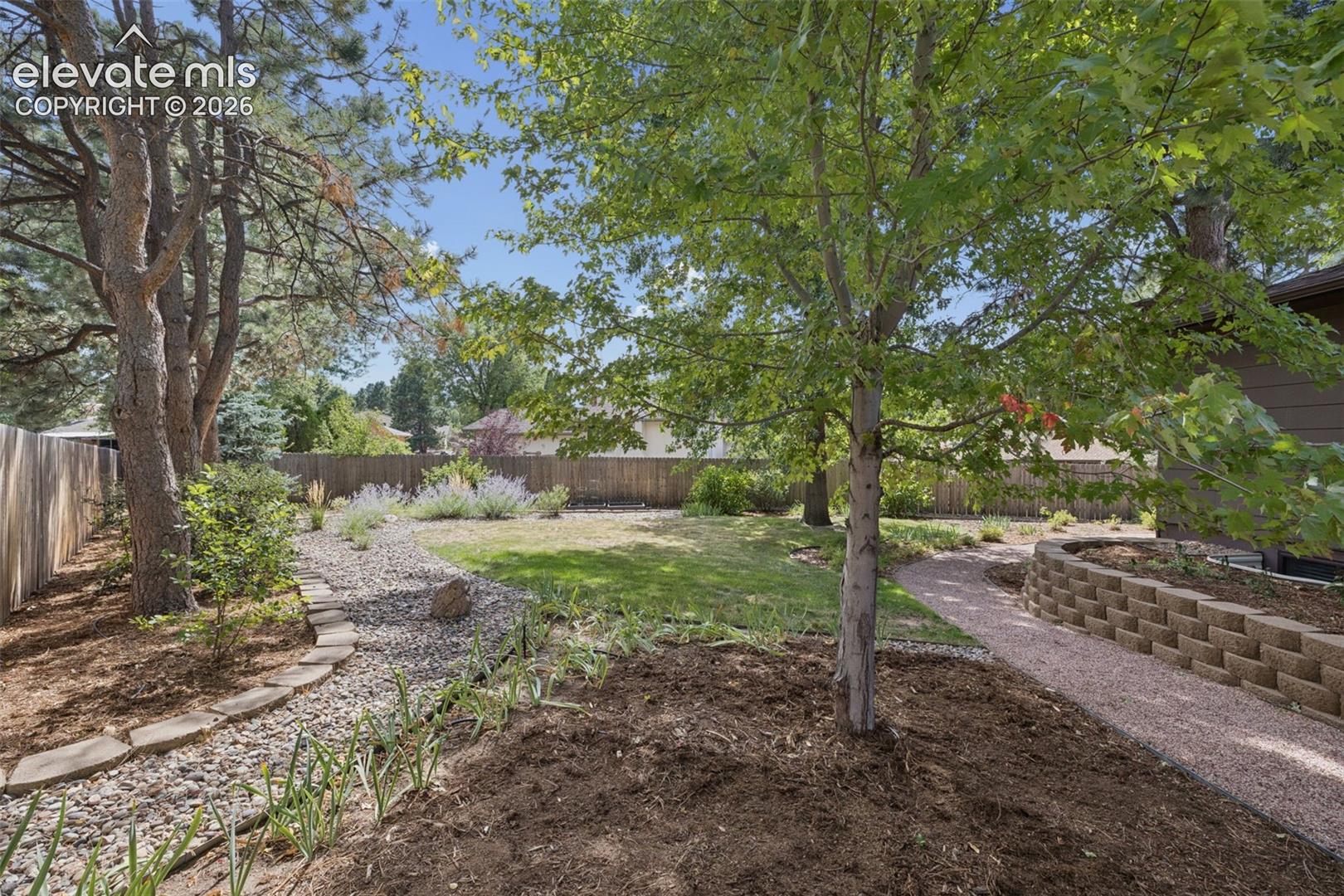 Backyard featuring a stone-retaining wall, gravel pathway, mature trees, tiered landscaping, and wood perimeter fencing