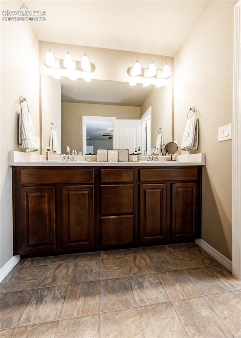 Dual-sink vanity with dark wood-finish cabinetry, solid surface countertop, two multi-bulb vanity lights, and tile flooring