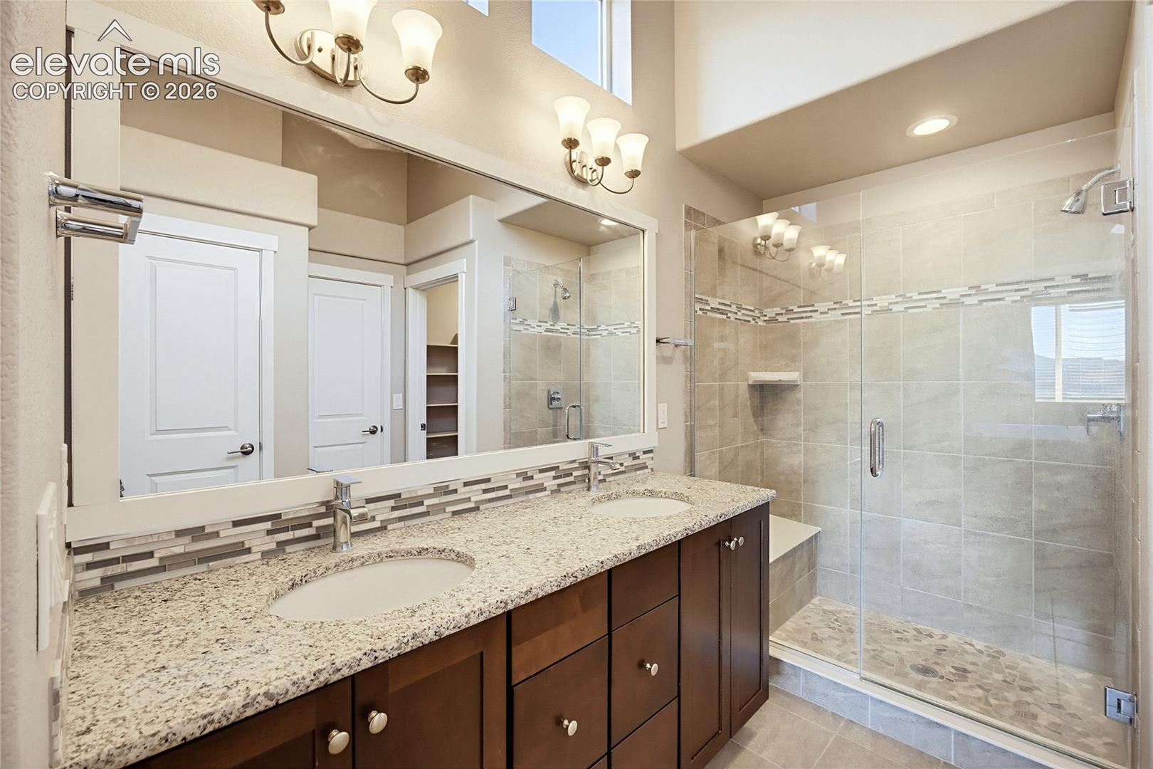 Dual-sink vanity with stone-finish countertop, full-width mirror, glass-enclosed shower featuring tiled surround, and integrated wall lighting