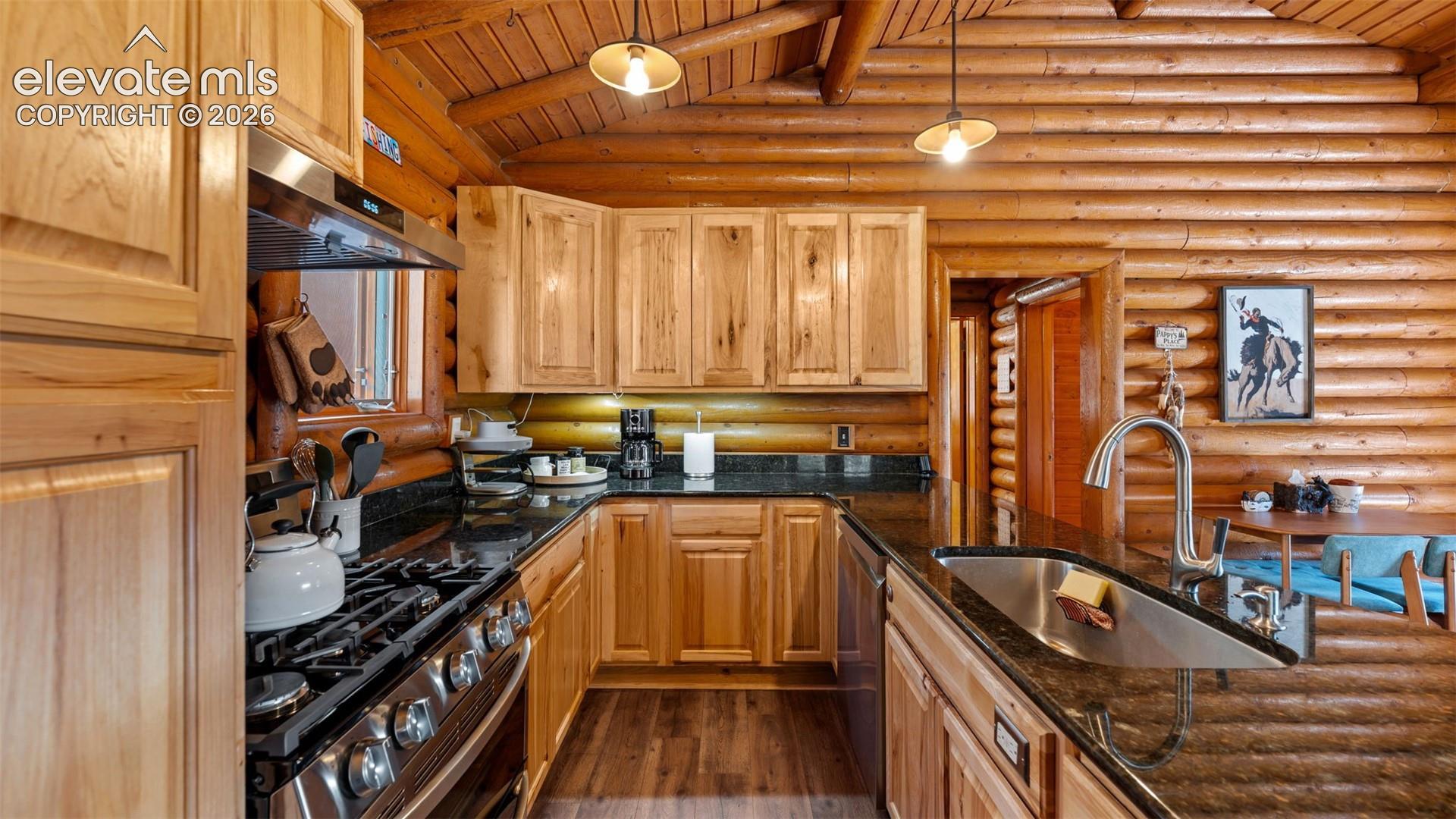 Kitchen featuring natural log walls, custom wood cabinetry, dark stone countertops, stainless steel gas range, and pendant lighting