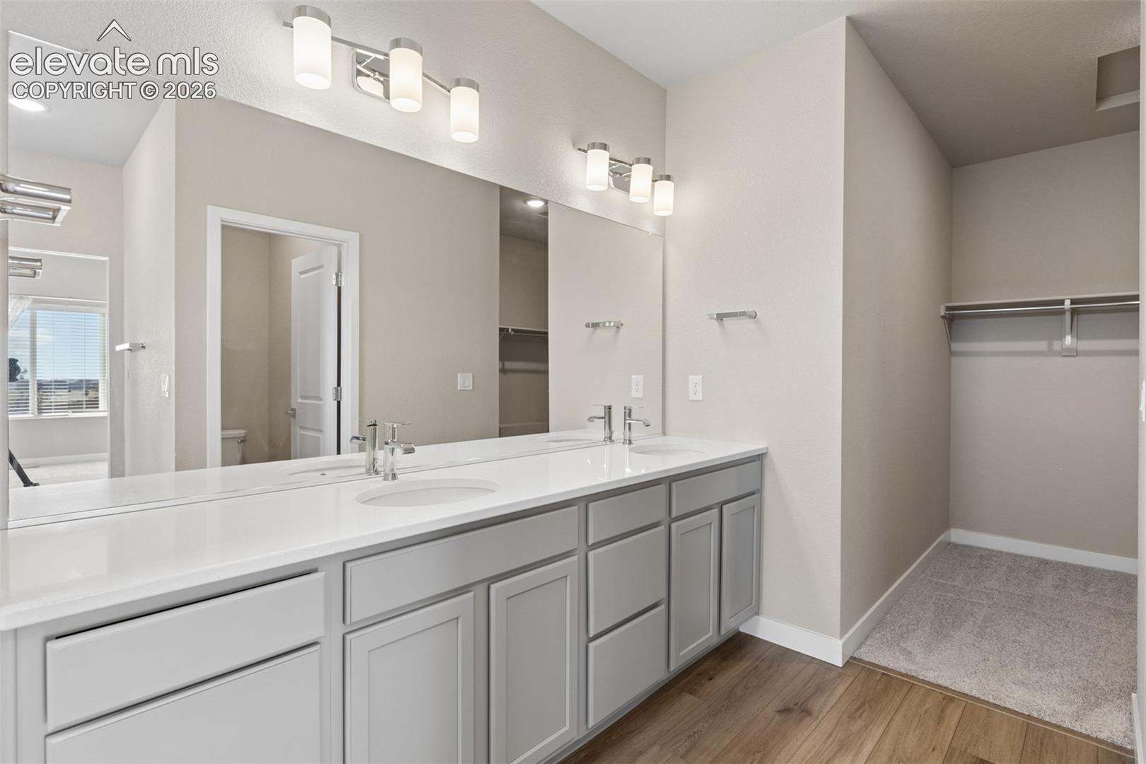 Dual-sink vanity with a solid-surface countertop, full-width wall mirror, double wall-mounted vanity lights, wood-finish flooring, and built-in closet shelving