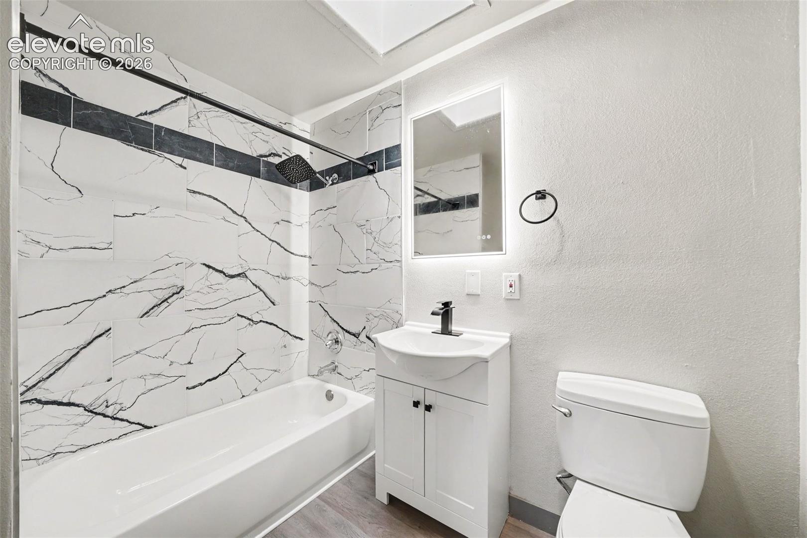Bathroom featuring a shower-tub combination with patterned tile surround, white vanity, vessel sink, matte black faucet, and backlit mirror