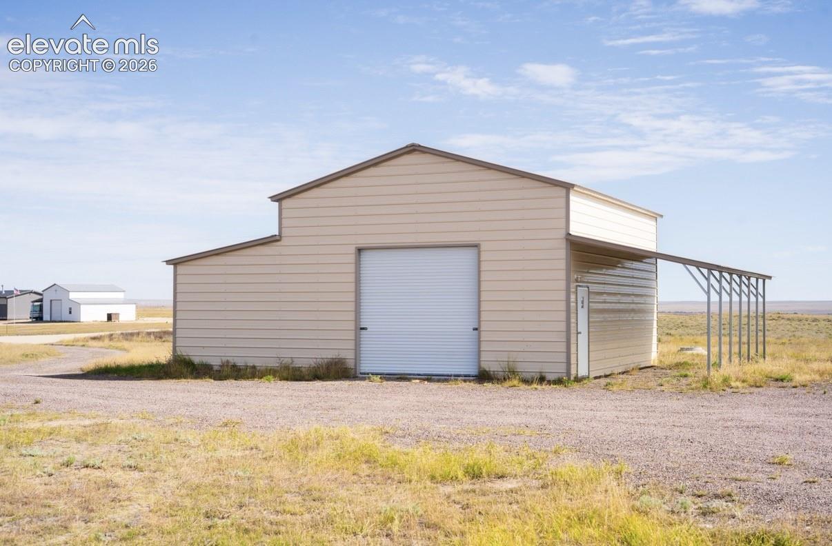 Detached metal workshop featuring beige horizontal siding, a central roll-up garage door, an integrated personnel door, and an open-sided lean-to shelter