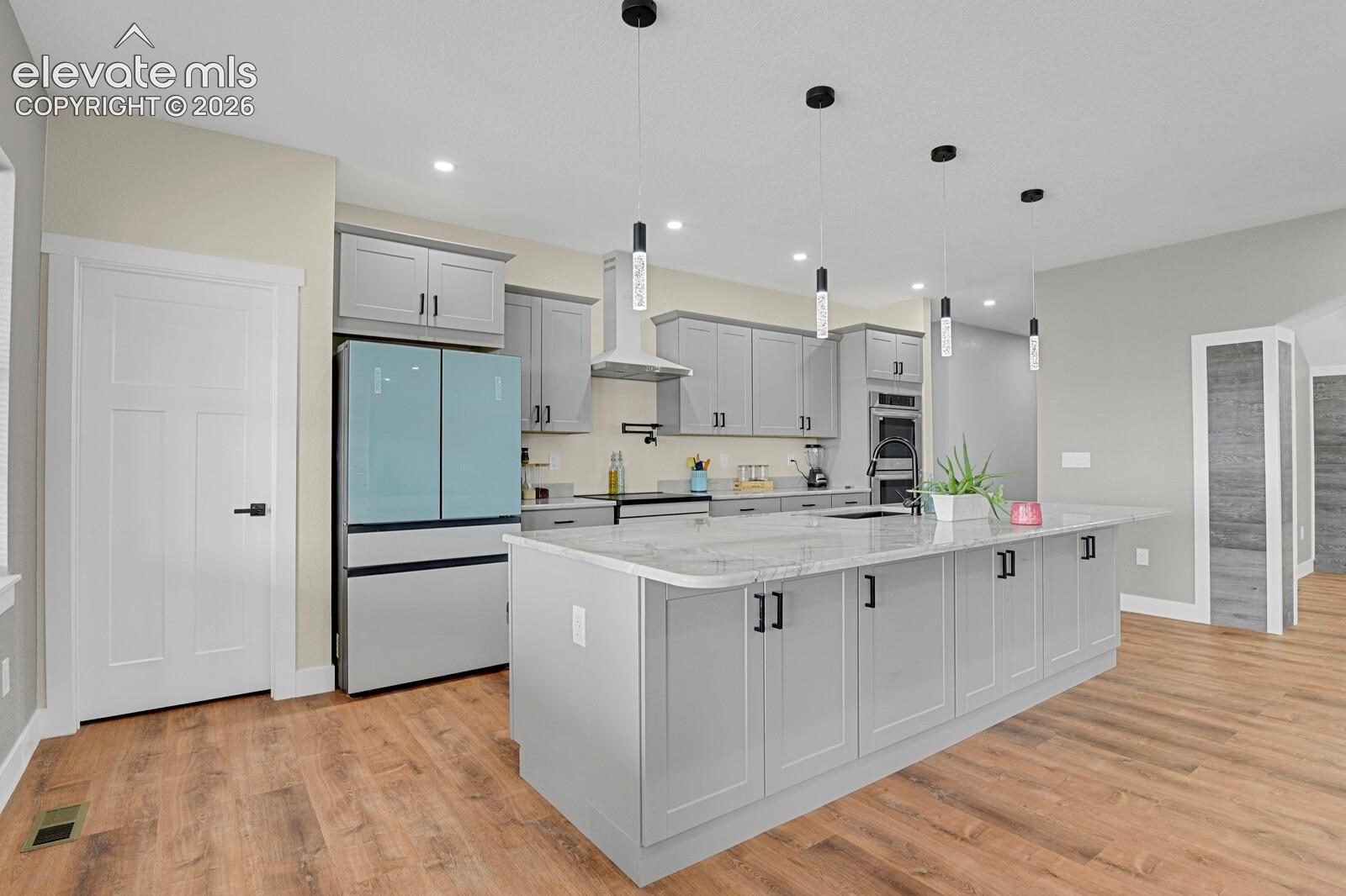 Spacious kitchen featuring an expansive island with a stone-finish countertop, light gray cabinetry with black hardware, wood-finish flooring, and pendant lighting