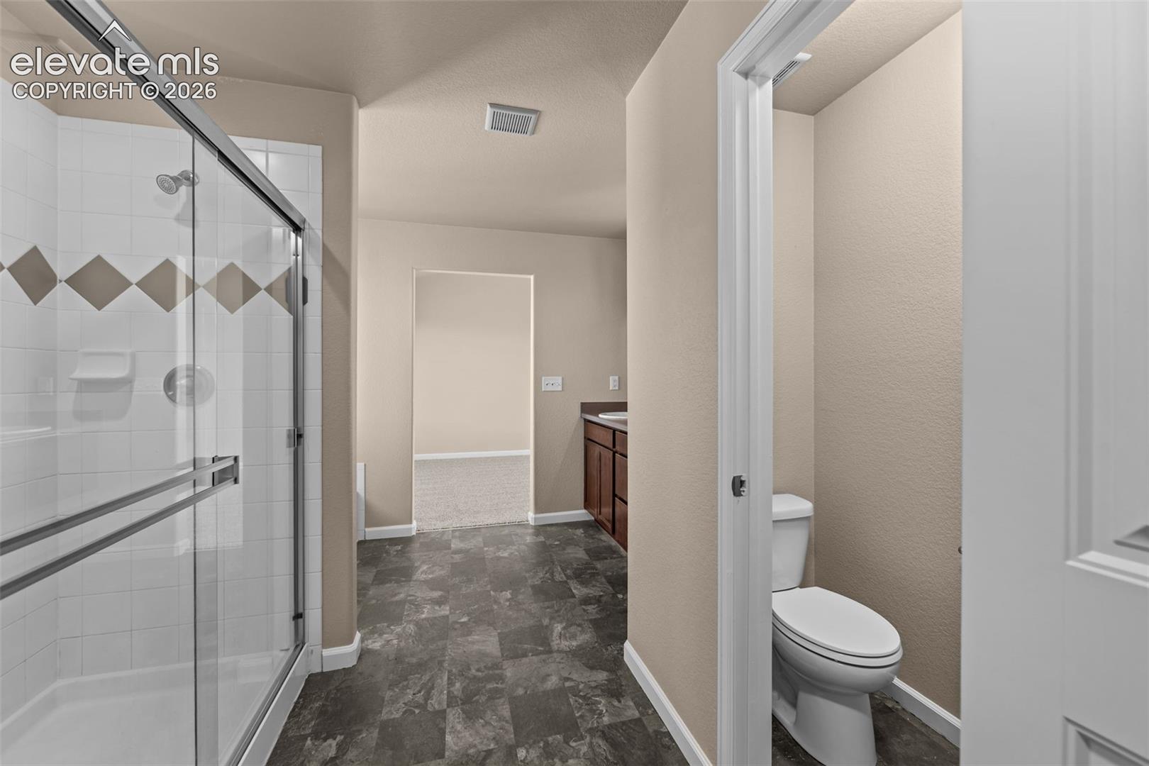 Bathroom featuring a glass-enclosed shower with tiled surround, wood-finish cabinetry, stone-patterned vinyl flooring, and a separate water closet
