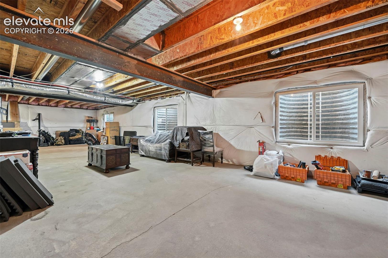 Exposed structural beams, two windows with window wells, concrete flooring, and insulated wall surfaces create an unfinished basement space
