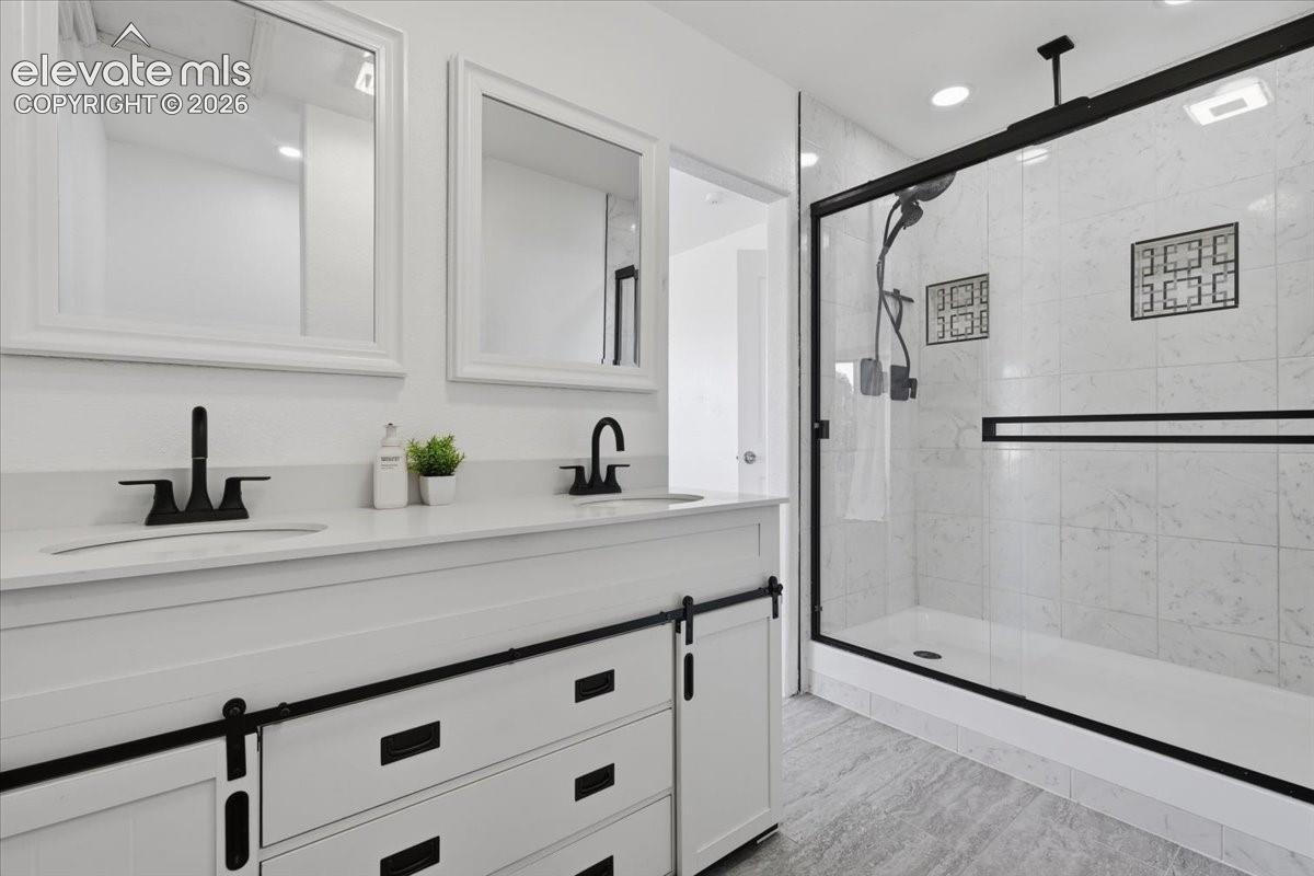Dual vanity featuring a white cabinet with sliding hardware, stone-surface countertop, matte black fixtures, framed mirrors, and a walk-in shower with stone-tile surround and a matte black sliding glass door