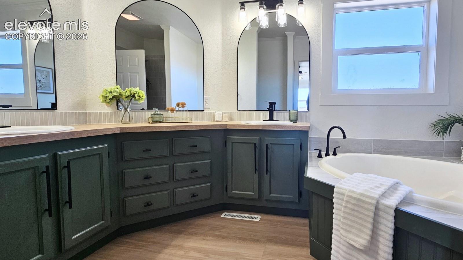 Dual vanity with green cabinetry, wood-surface countertop, arched mirrors, matte black hardware, and an integrated soaking tub