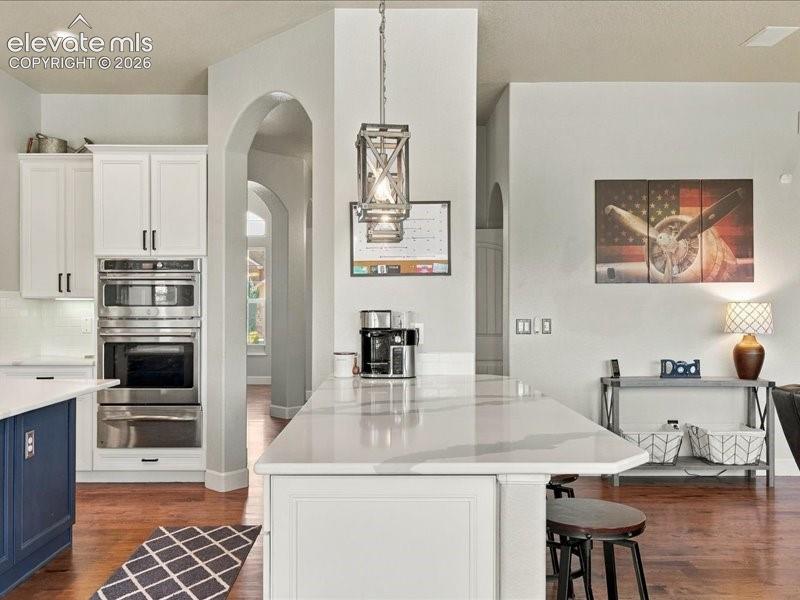 Kitchen featuring a spacious island with a light-toned countertop, white cabinetry with dark hardware, integrated stainless steel double ovens, and wood-finish flooring