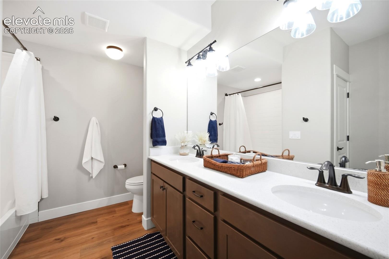 Bathroom featuring a dual-sink vanity with light-toned solid surface countertops, dark-finish hardware, wood-finish flooring, and a full-width mirror