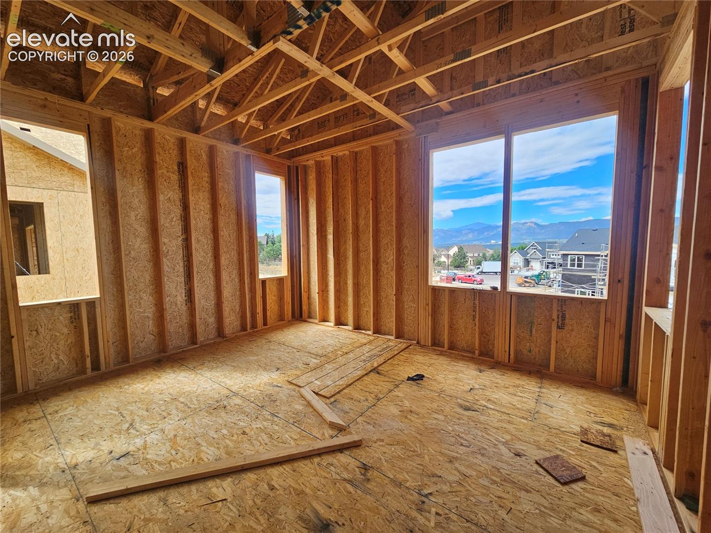 other room under construction with exposed wood stud framing, roof trusses, and large windows showing neighborhood and distant mountains