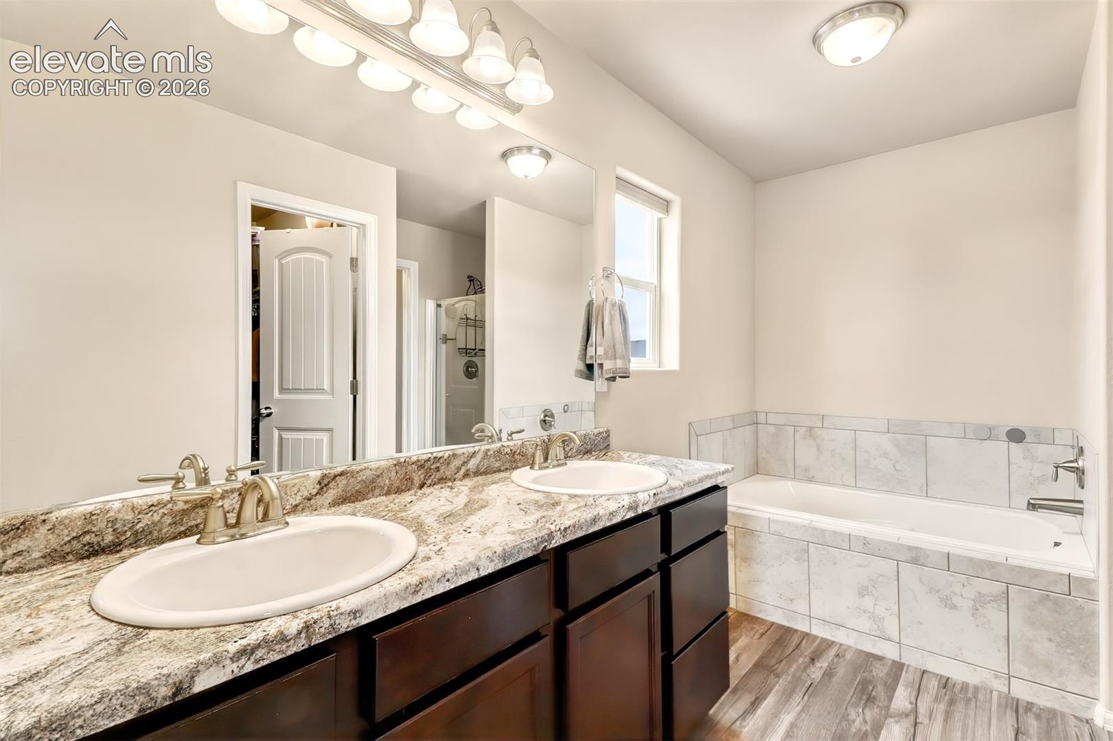 Dual-sink vanity with stone-patterned countertops, dark wood cabinetry, wood-finish flooring, and a tiled soaking tub surround