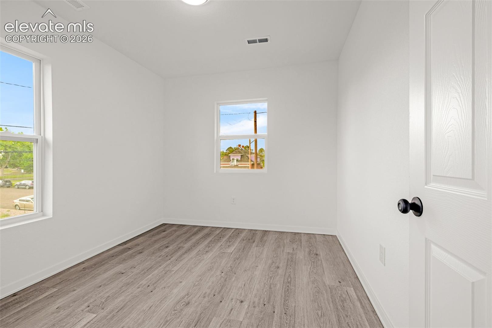 Room featuring wood-finish flooring, two windows, white walls, and a white door with black hardware