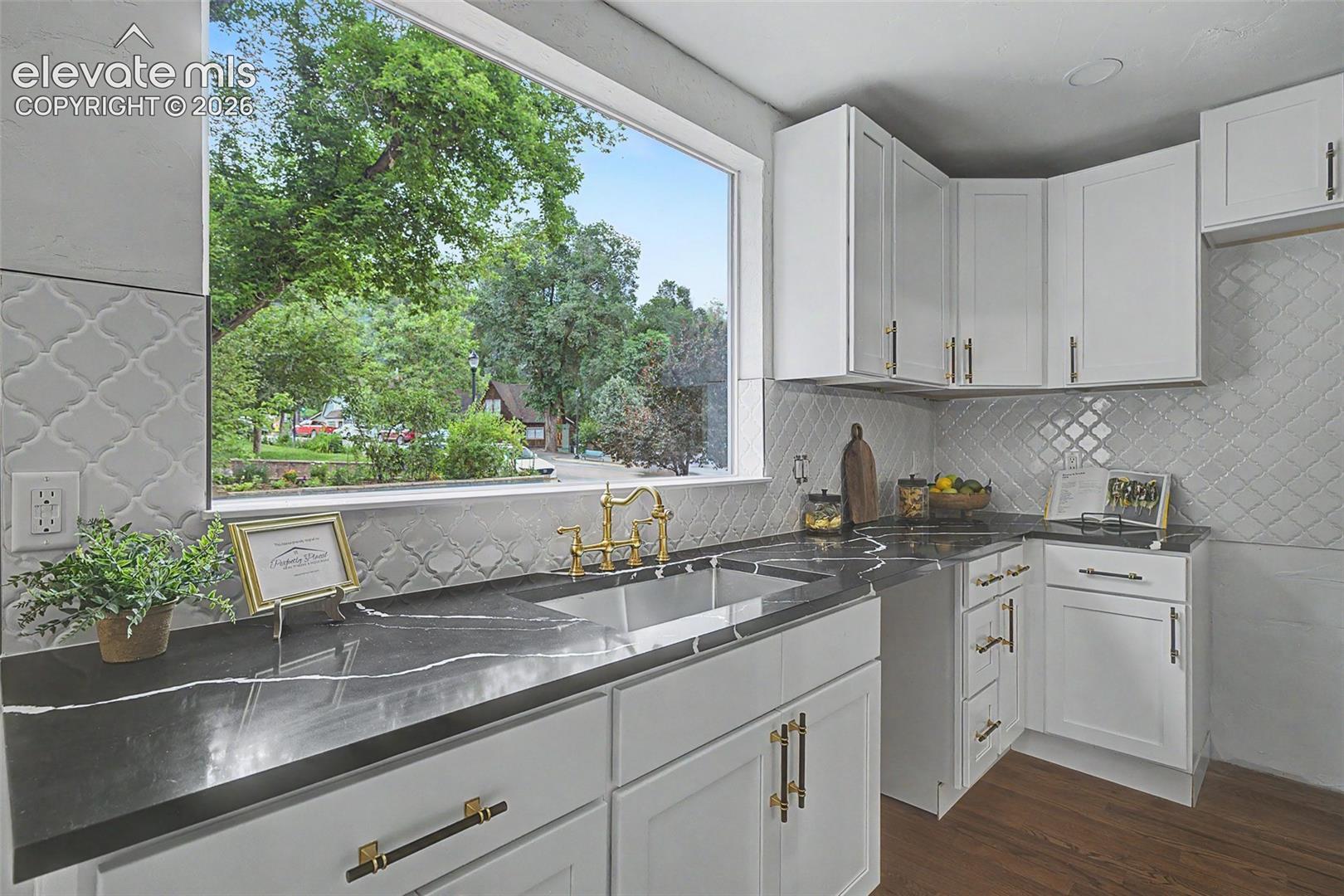 Kitchen featuring white shaker-style cabinetry, dark countertops with light veining, an arabesque-patterned backsplash, a large single-basin sink, and brass-tone hardware