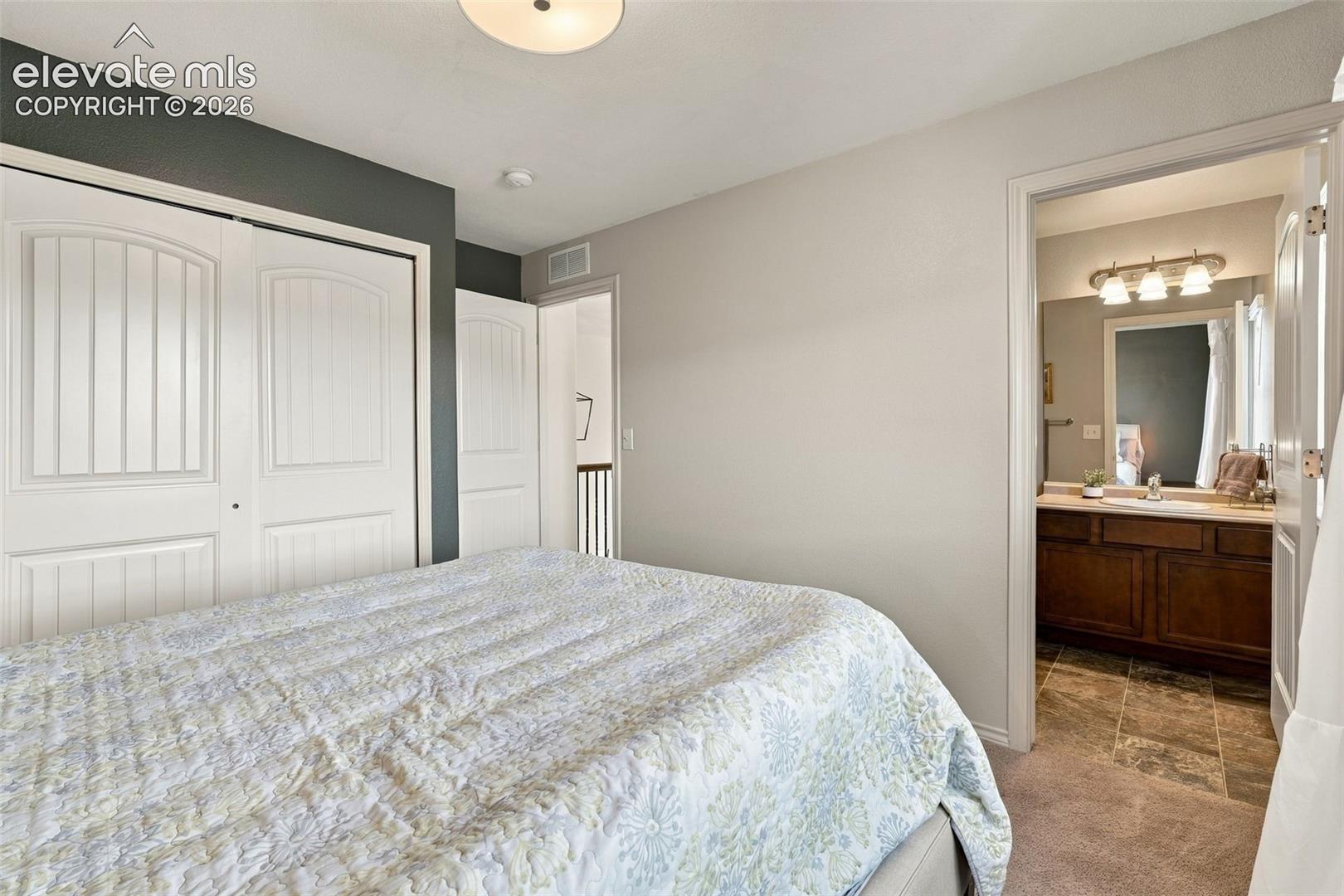 Bedroom featuring double-door closets and neutral carpeting, with an attached bathroom containing a wood-finish vanity and stone-patterned tile flooring