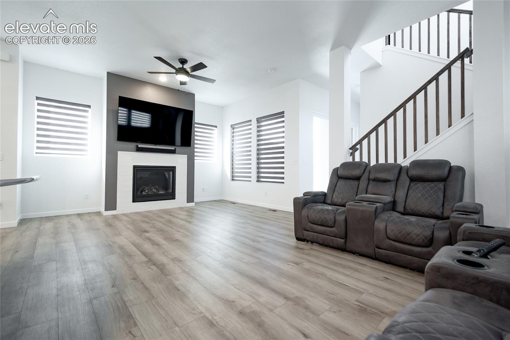 Open living area featuring wood-finish flooring, a fireplace with a tiled surround, a ceiling fan, dual-tone accent wall, and a staircase with a wooden railing