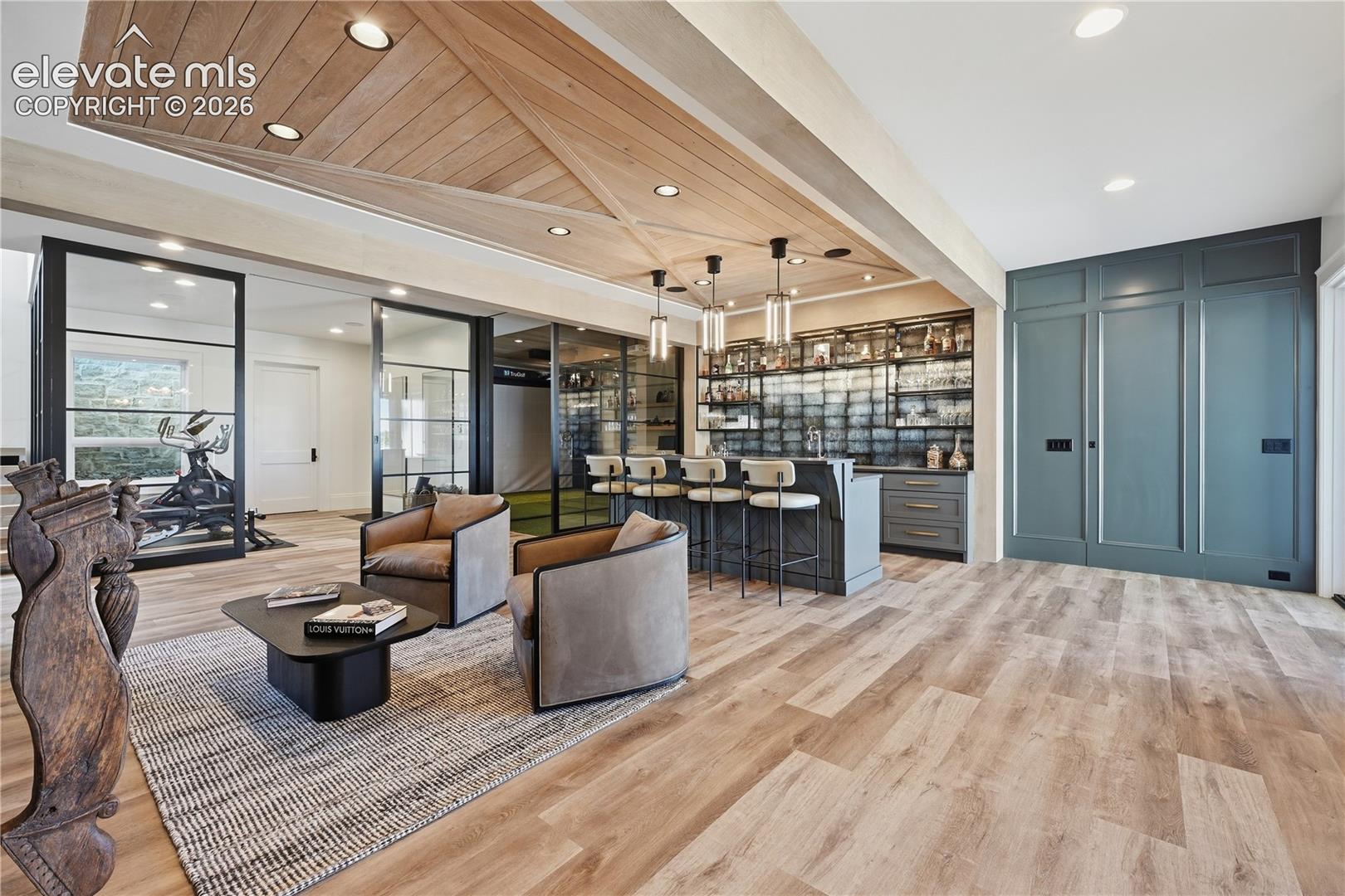 Custom wood-paneled ceiling feature with recessed lighting, integrated wet bar with glass-front shelving, full-height paneled wall cabinetry, and glass-paneled interior partitions with metal framing