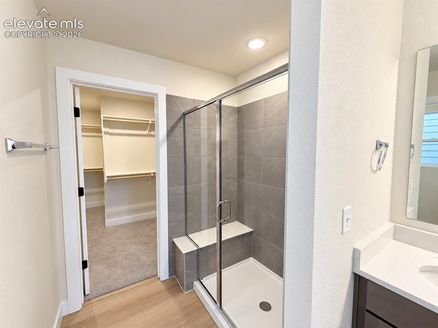 Walk-in shower featuring gray tile surround, built-in bench seat, and glass enclosure, plus built-in closet shelving and vanity with solid-surface countertop