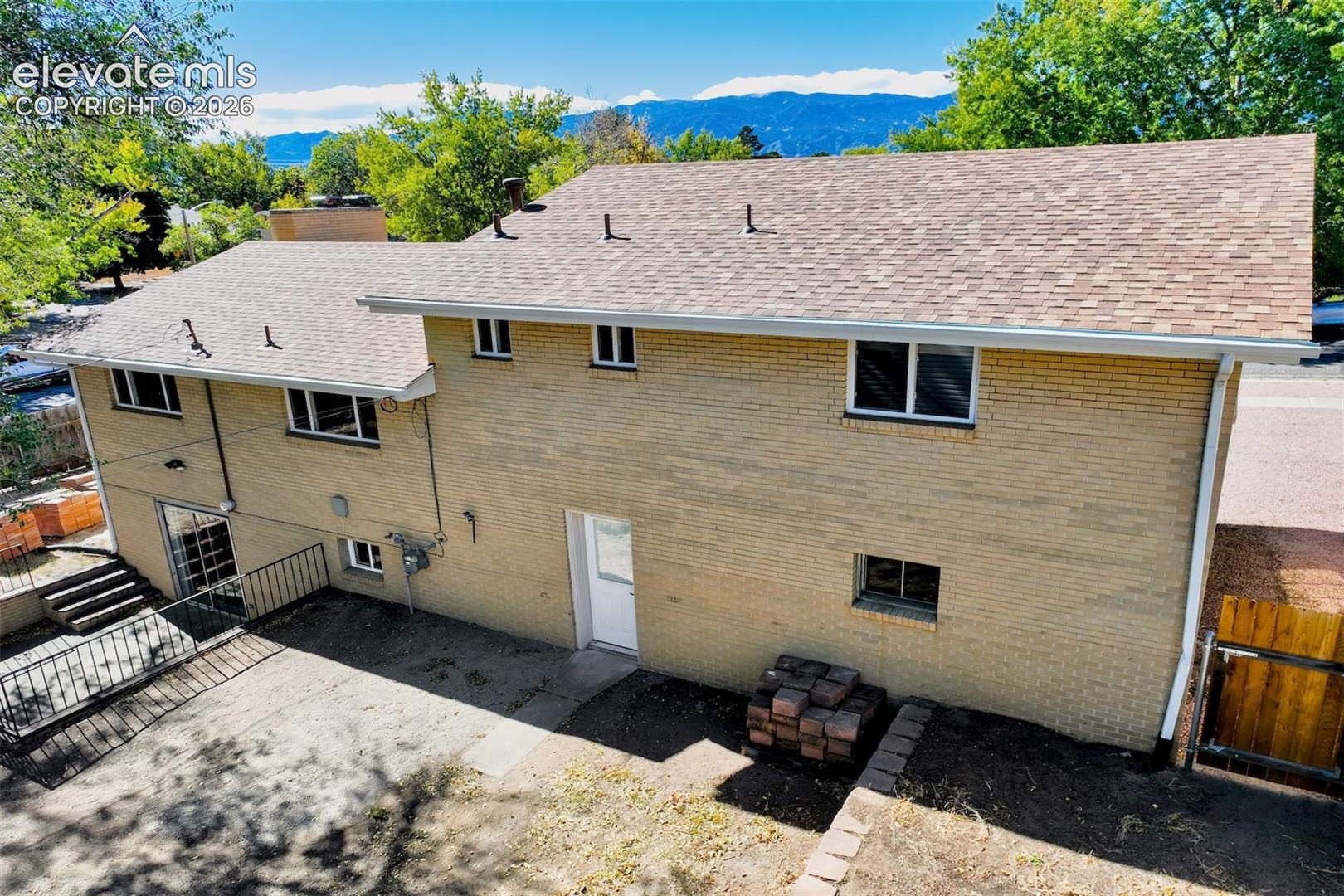Multi-level brick exterior with composition shingle roofing, concrete patio, metal railings, and mountain backdrop